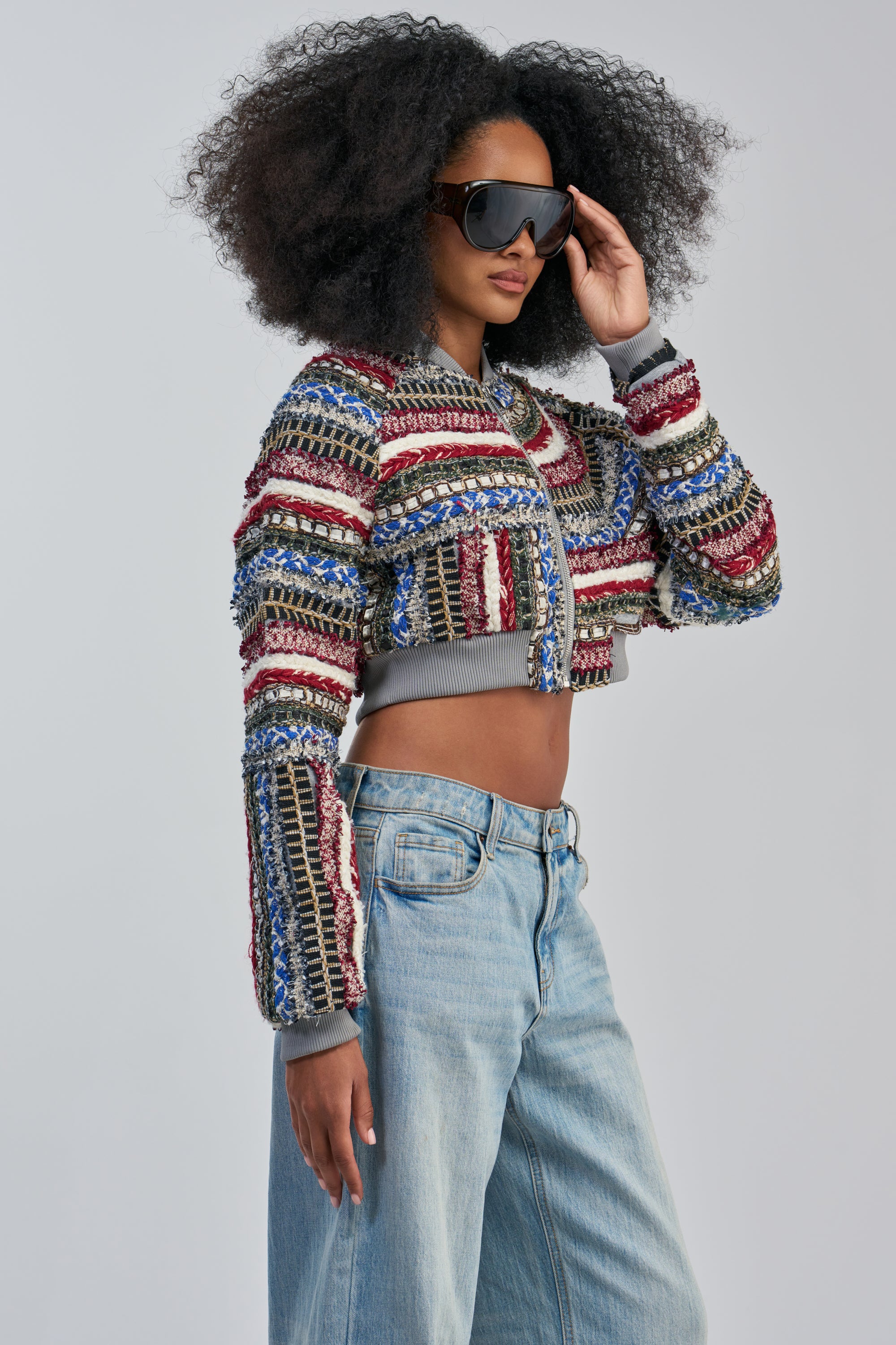 Wearing the FAYE TEXTURED BOMBER—a vibrant, multicolor jacket with textured details—a Black woman with voluminous curly hair and oversized sunglasses poses against a plain grey background, her hand resting on the rim of her glasses. She completes the look with light-wash blue jeans.