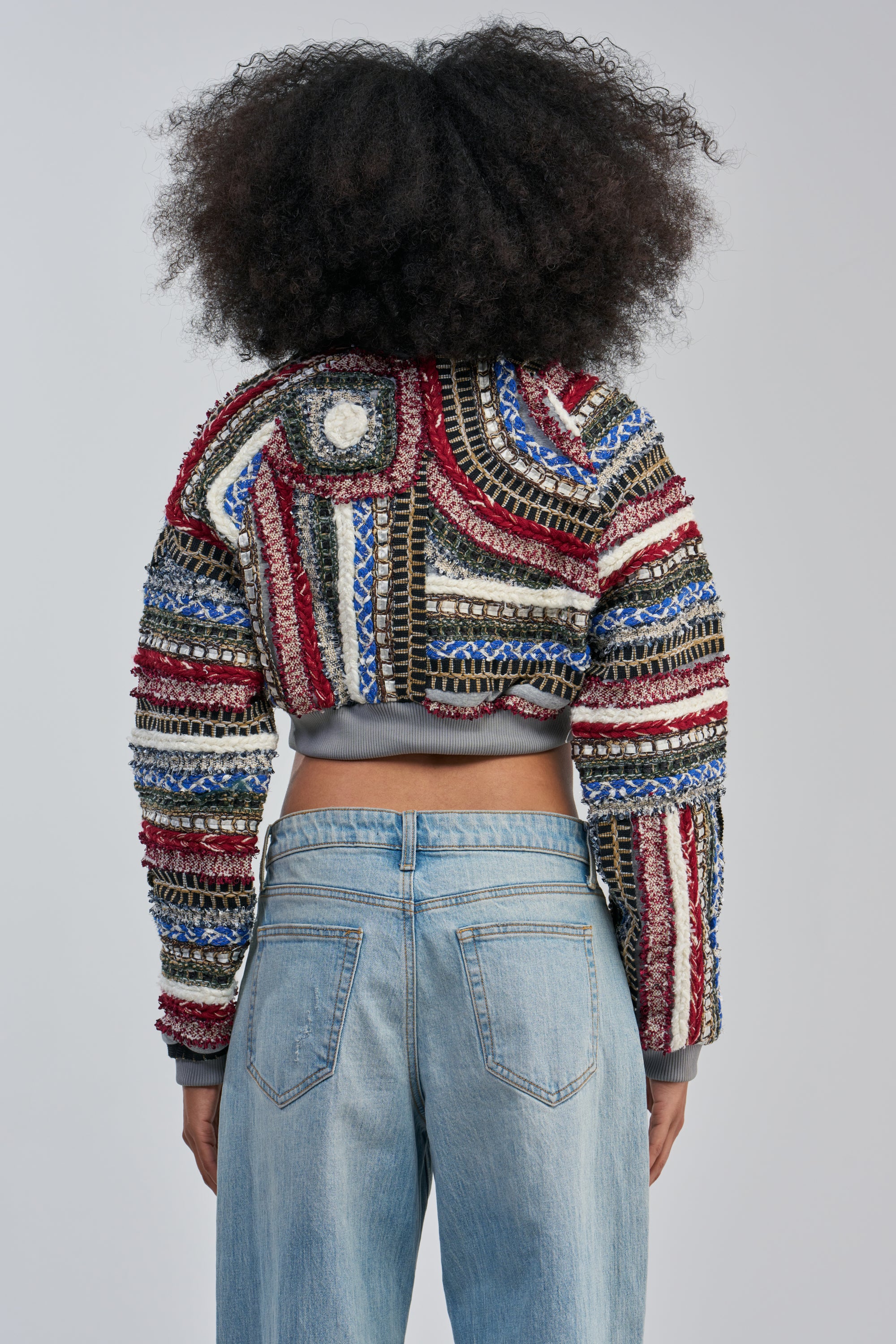 A person with voluminous, curly hair is photographed from behind against a plain light gray background, wearing the FAYE TEXTURED BOMBER—a colorful streetwear jacket featuring red, blue, white, and metallic textured patterns—paired with light blue jeans.