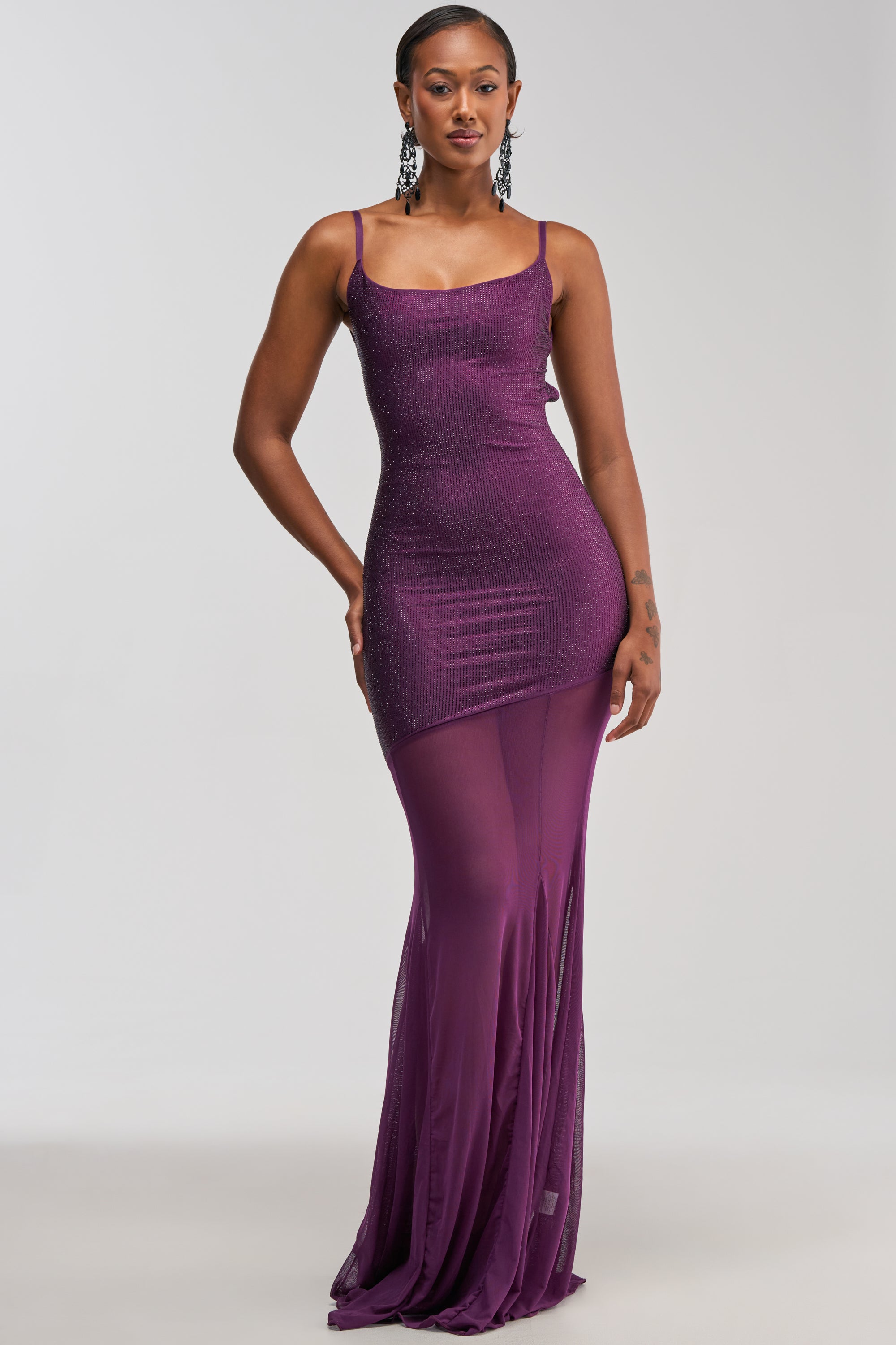 Confidently standing against a plain light gray background, the woman is wearing the SHOW A LITTLE LEG RHINESTONE MAXI DRESS IN PLUM—a sleeveless, form-fitting, glamorous evening gown featuring glittery rhinestone accents in a rich plum hue and a sheer lower half.