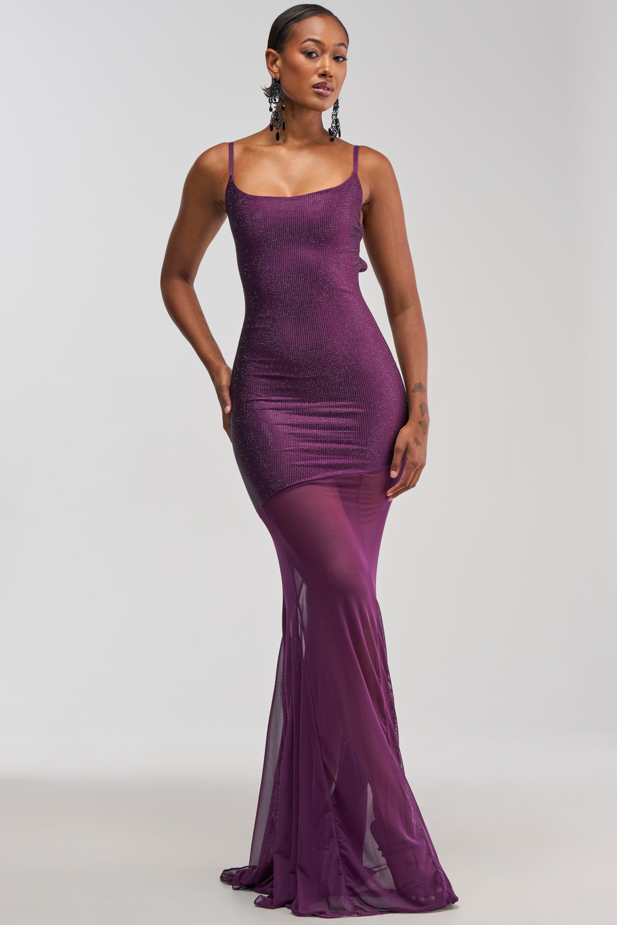 A woman poses in the SHOW A LITTLE LEG RHINESTONE MAXI DRESS IN PLUM—a sleeveless, fitted purple gown with a sheer, flowing lower half. She accessorizes with large black earrings and has short hair, standing against a simple light background.