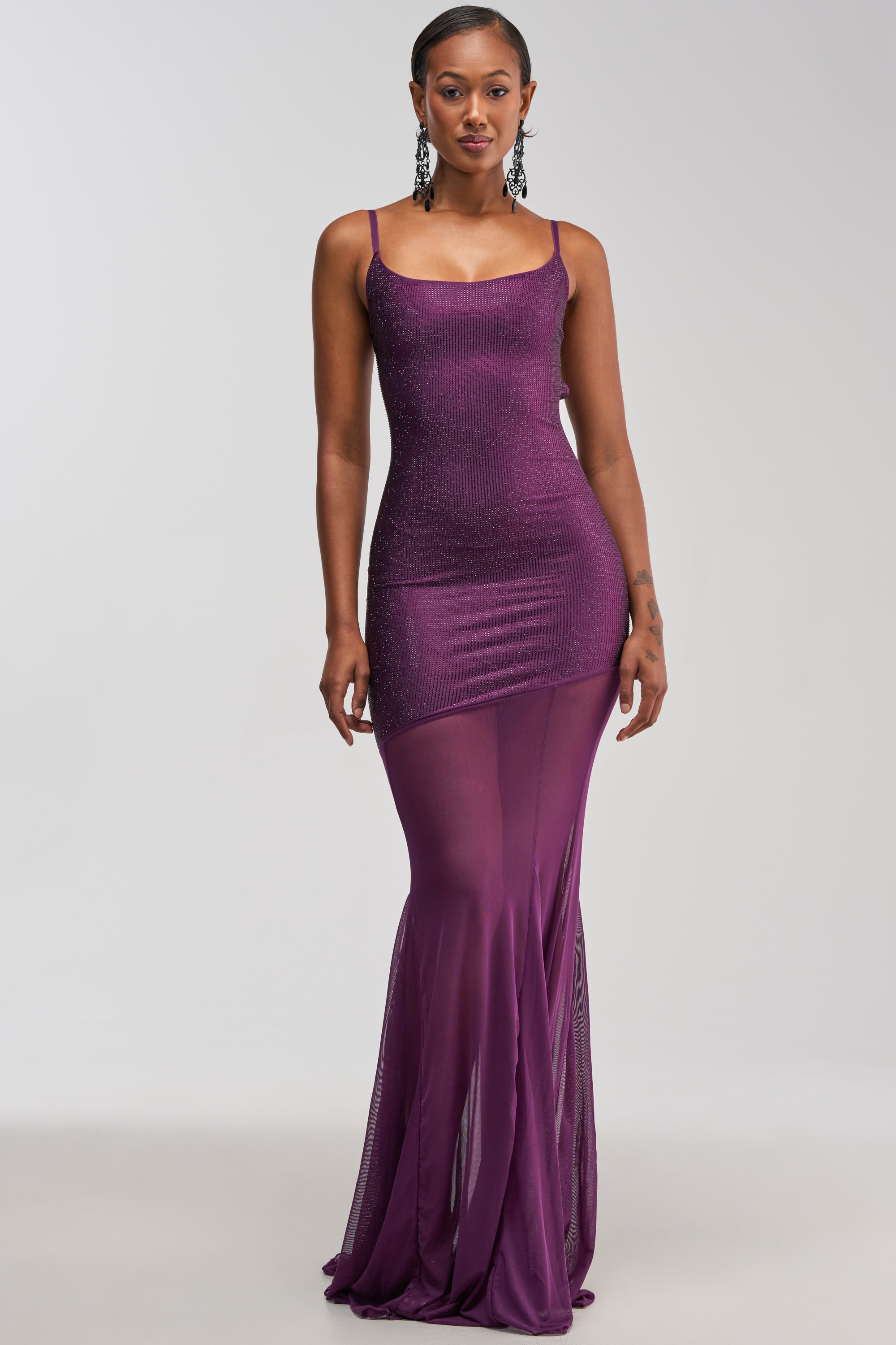 A woman poses against a plain light background, wearing the SHOW A LITTLE LEG RHINESTONE MAXI DRESS IN PLUM—a fitted, sleeveless, floor-length purple dress with a sheer skirt and thin straps. She completes her look with statement earrings and short, slicked-back hair.