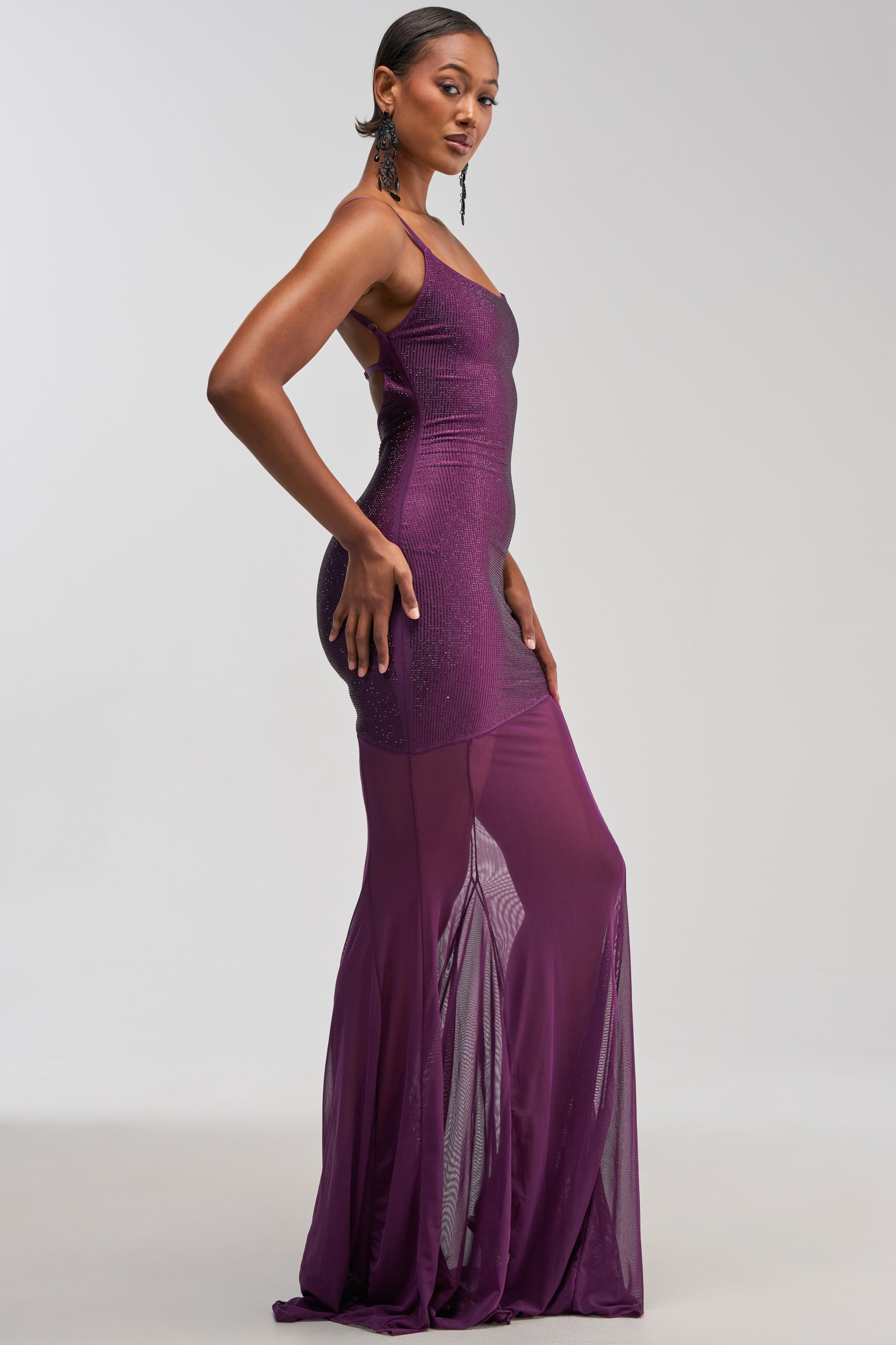 A woman with short hair, accessorized with long, dangling earrings, poses sideways against a plain light background wearing the SHOW A LITTLE LEG RHINESTONE MAXI DRESS IN PLUM—a fitted, sleeveless maxi dress featuring sparkling rhinestones and sheer lower panels.
