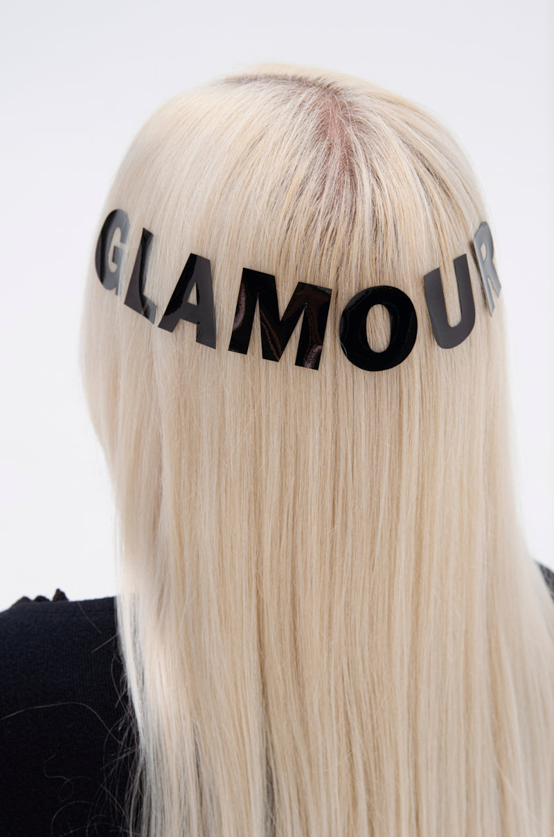 A person with long, straight platinum blonde hair wears a black headband with bold "GLAMOUR" letters and a GLAMOUR BODY AND HAIR STICKER, seen from behind against a plain light background. 0