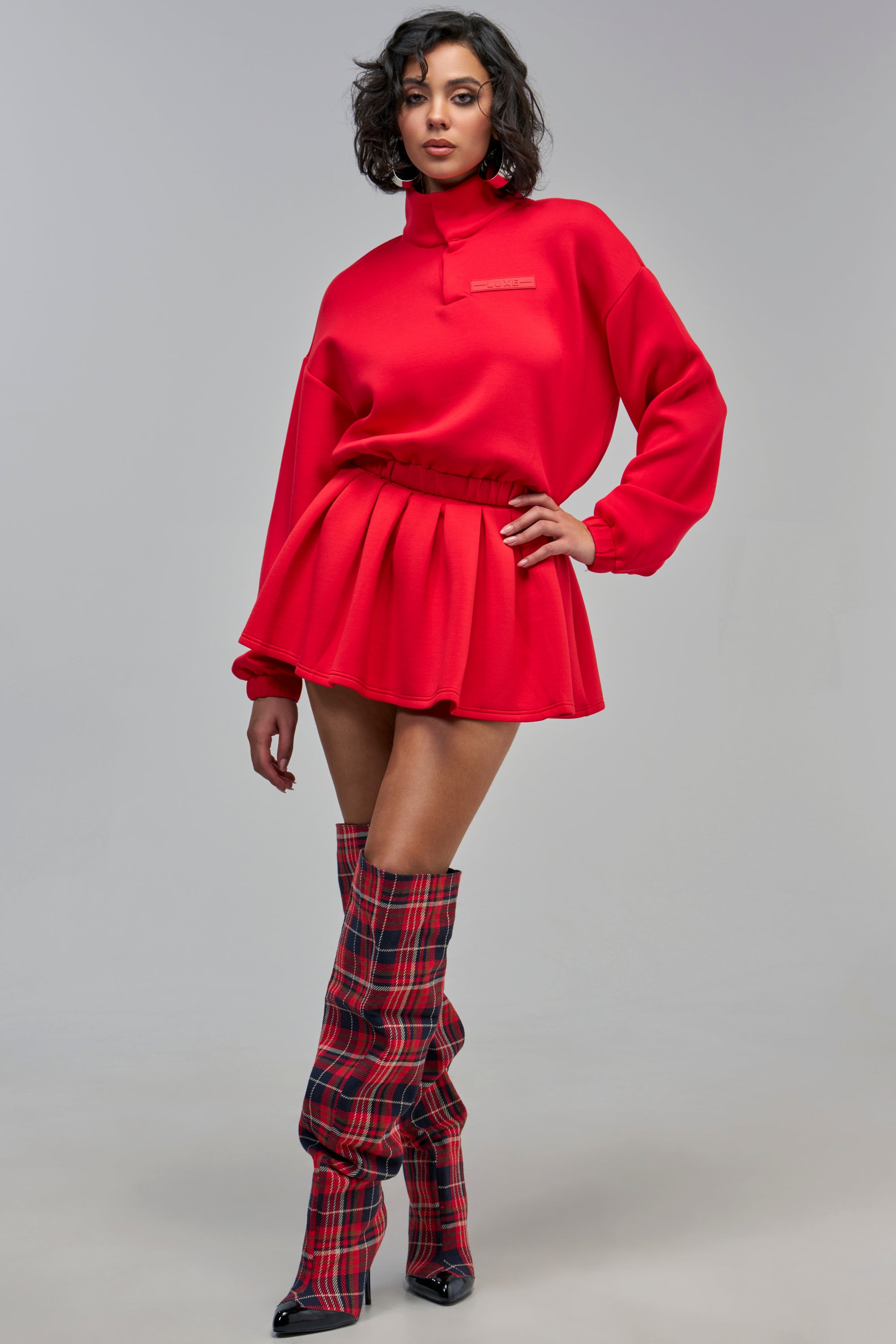 A person wears a red long-sleeved mock-neck sweatshirt tucked into a matching red PAXTON LUXE EVERYDAY ICON MINI SKIRT, paired with red plaid over-the-knee boots in a preppy loungewear style.