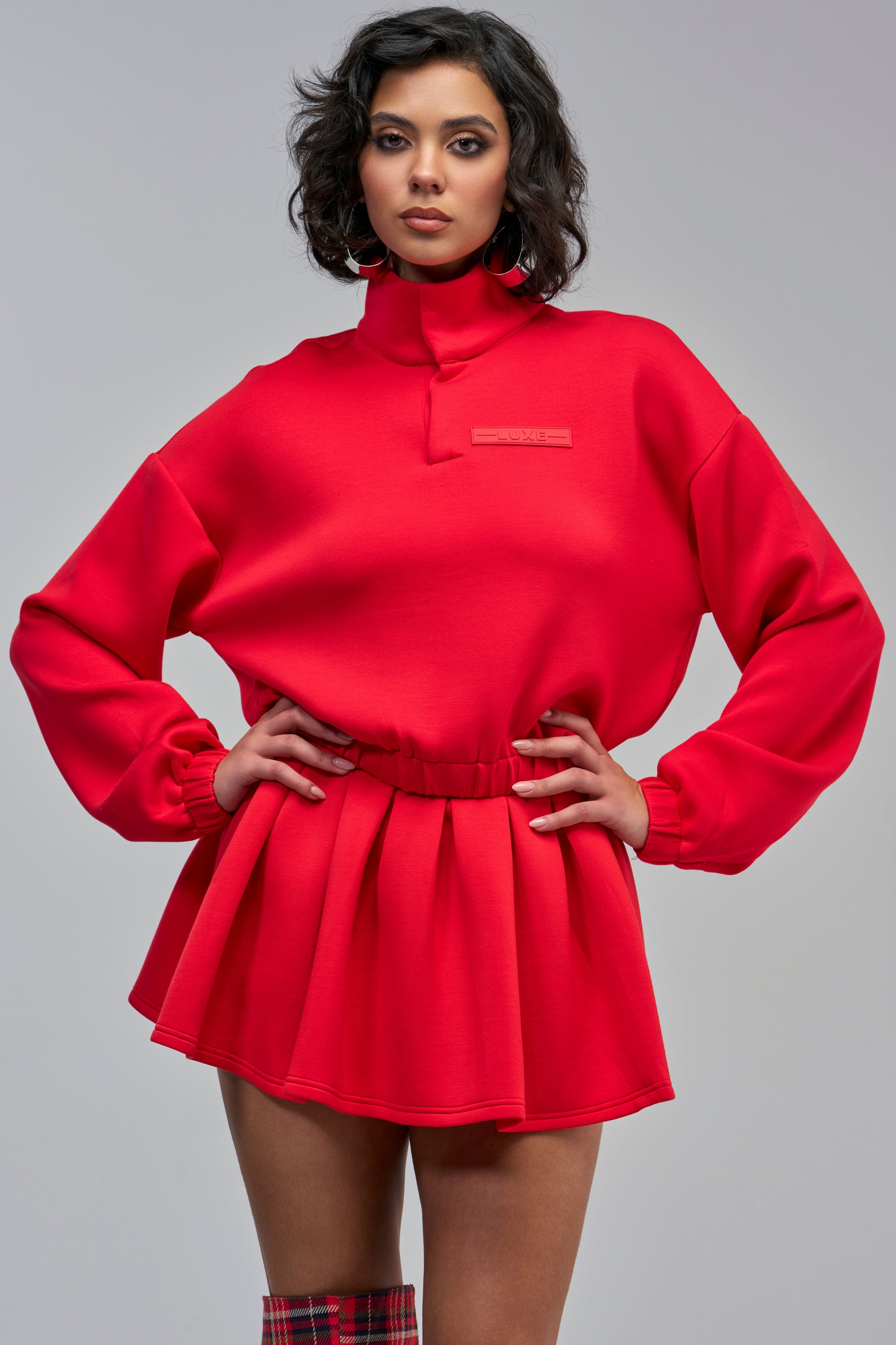 A person wearing a bright red PAXTON LUXE EVERYDAY ICON QUARTER ZIP SWEATSHIRT, a cropped style featuring a pleated hem and a small “LUXE” label on the chest.