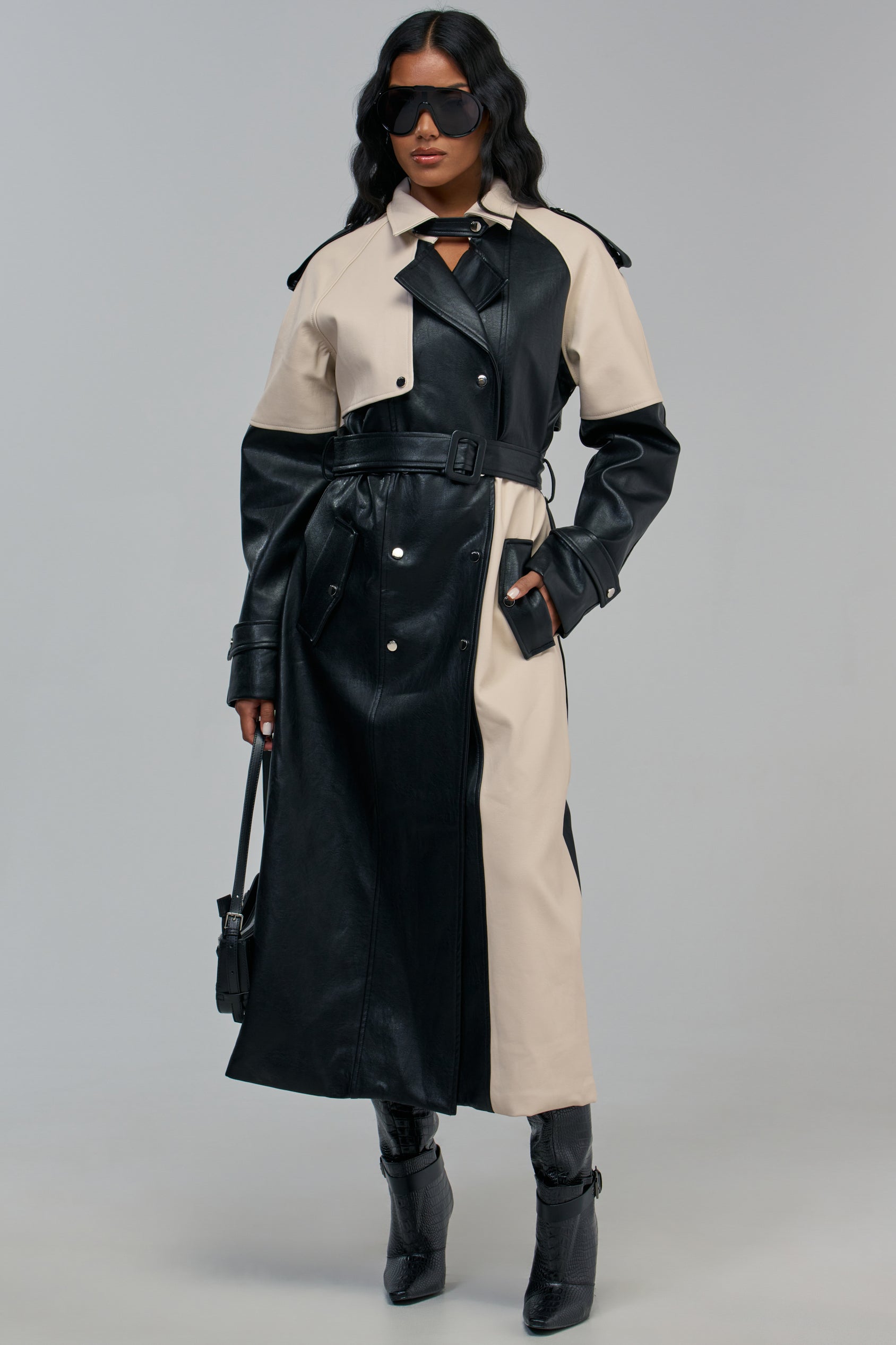 A person wears a LEX COLORBLOCKED TRENCH in black and beige with a belted waist, dark sunglasses, and black ankle boots.