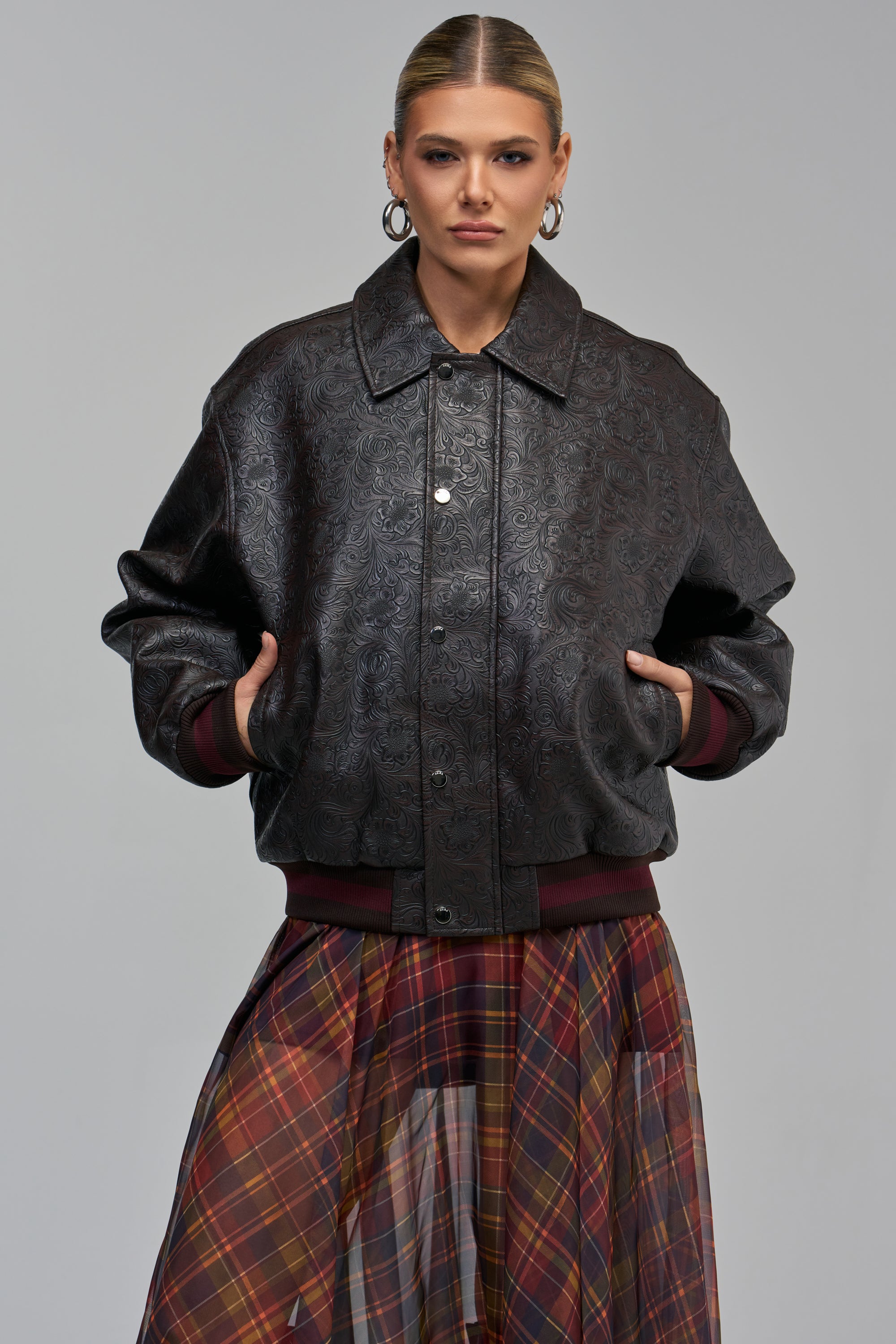 A person wears a dark floral UP CLOSE EMBOSSED BOMBER jacket over a sheer plaid skirt.