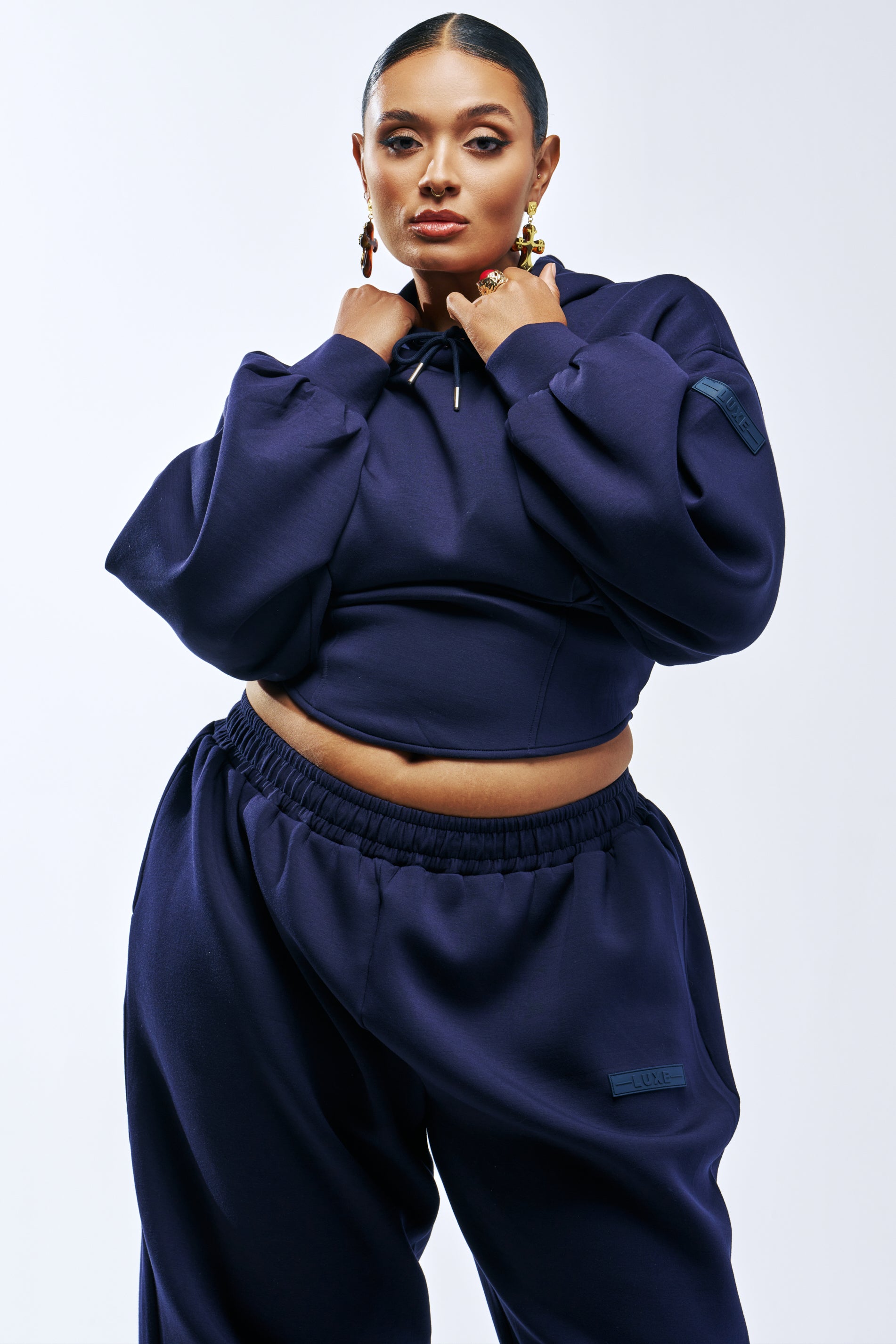A person wears a navy blue PAXTON LUXE MIDNIGHT SCULPT HOODIE with a corseted design and matching high-waisted sweatpants.