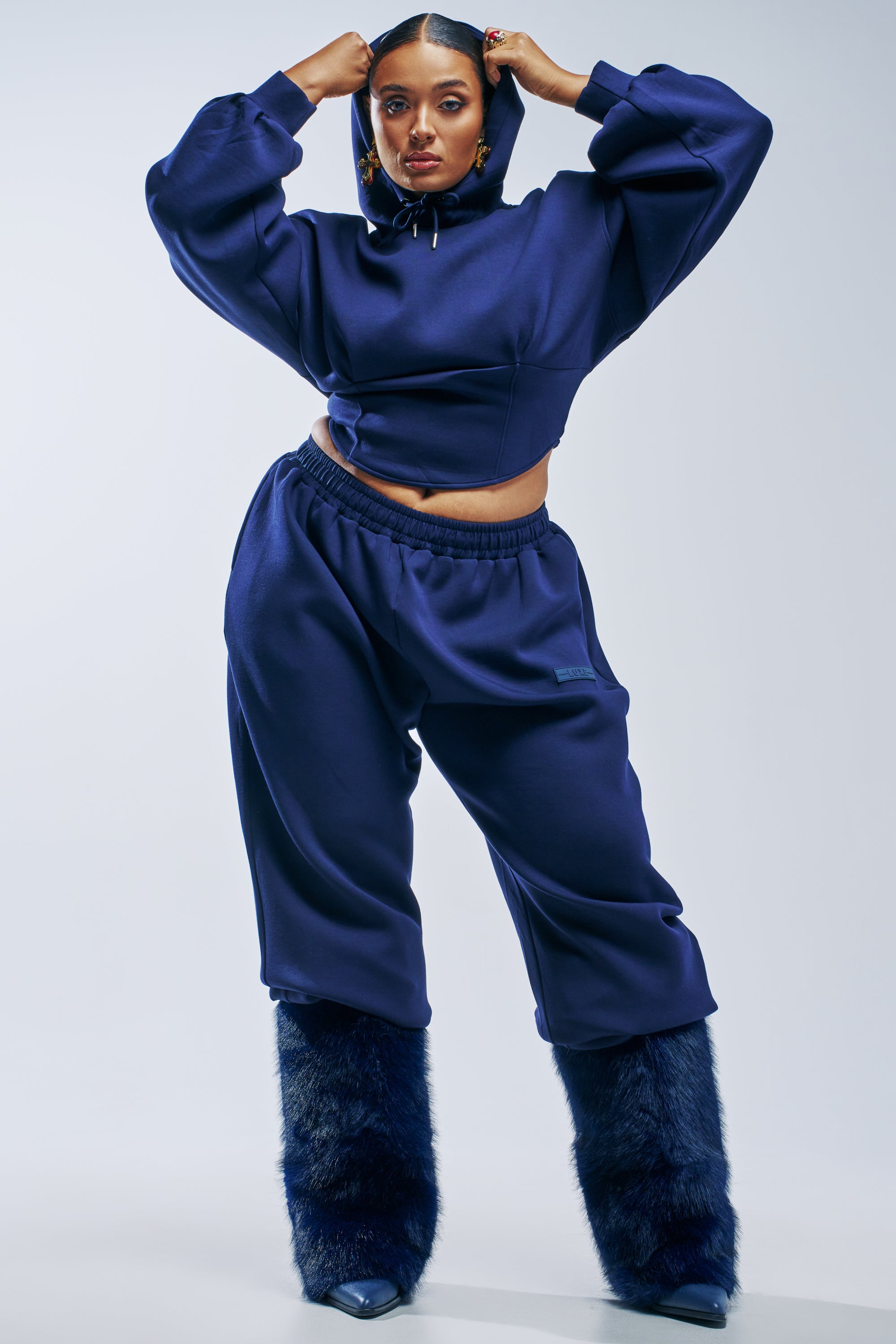 A person wears a dark blue cropped hoodie and matching PAXTON LUXE MIDNIGHT SCULPT JOGGER wide-leg joggers, paired with dark blue faux-fur leg warmers.
