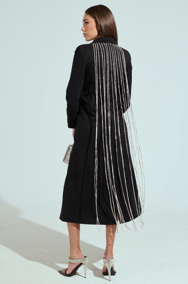 Against a light background, a woman poses sideways in the PARTY IN THE BACK RHINESTONE FRINGE TRENCH, its elegant longline rhinestone fringe falling from shoulders to floor. She completes the look with a glittery clutch and silver high heels.