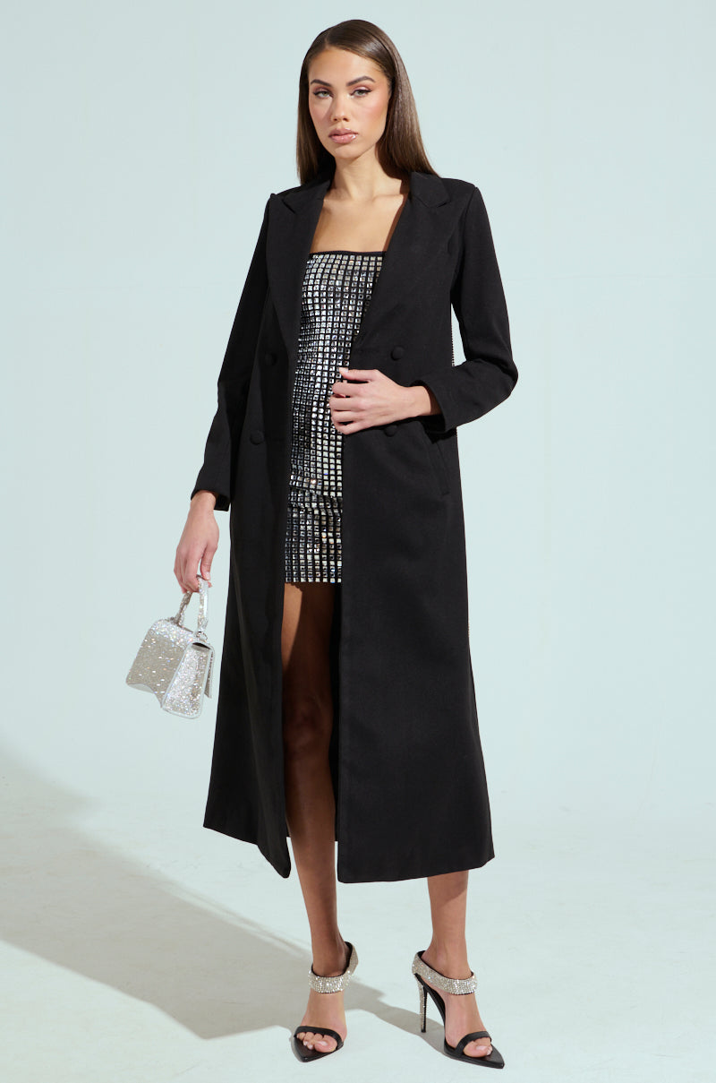 A woman models a longline silhouette wearing the PARTY IN THE BACK RHINESTONE FRINGE TRENCH, featuring a notch lapel collar over a sequin mini dress. She carries a sparkly handbag, wears high-heeled sandals, and poses with cascading hair against a light-colored background.