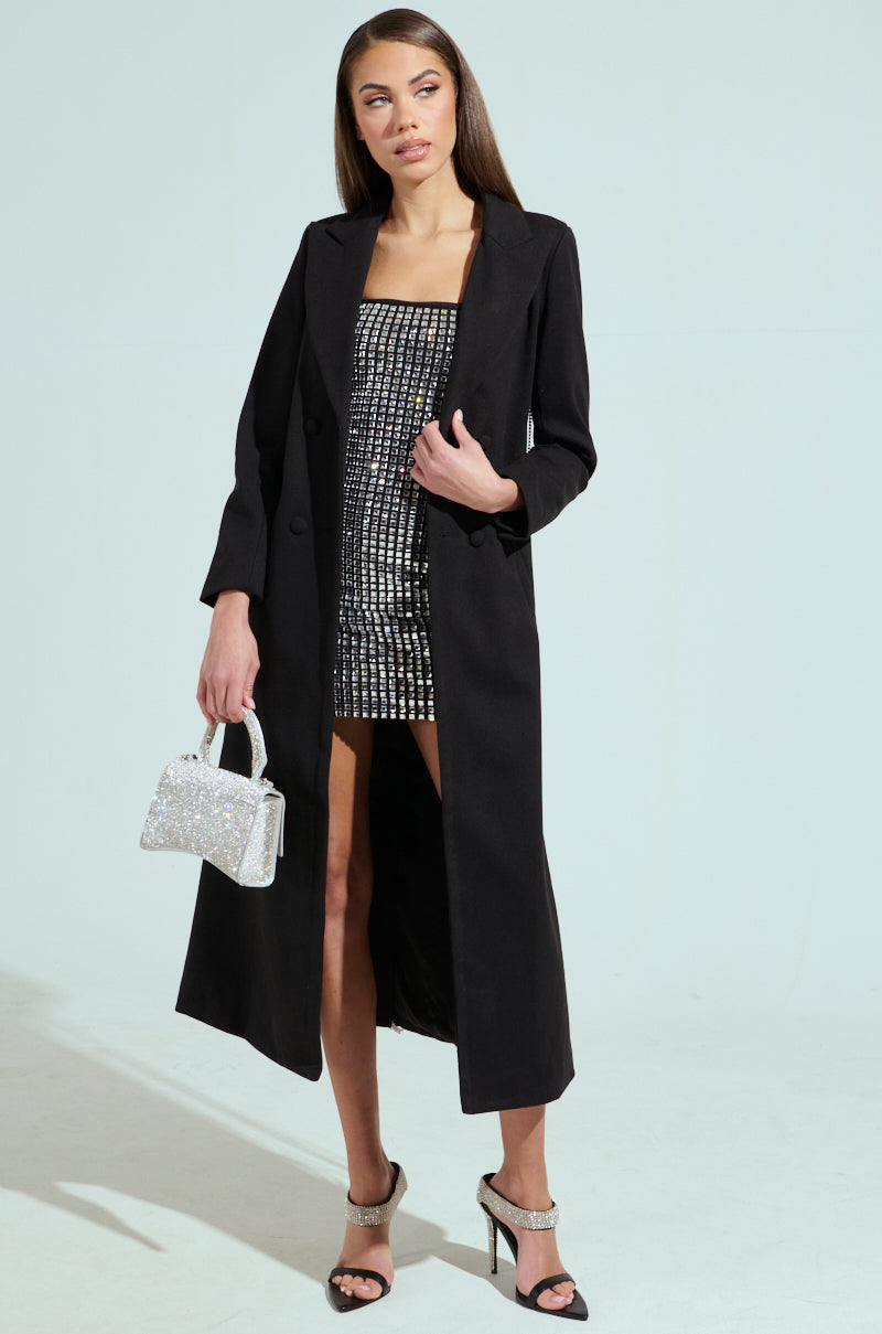 Draped in the Party in the Back Rhinestone Fringe Trench—a long black coat with a notch lapel collar—over a silver and black patterned dress, accessorized with a sparkling silver handbag and high-heeled strappy sandals, all set against a light-colored background.