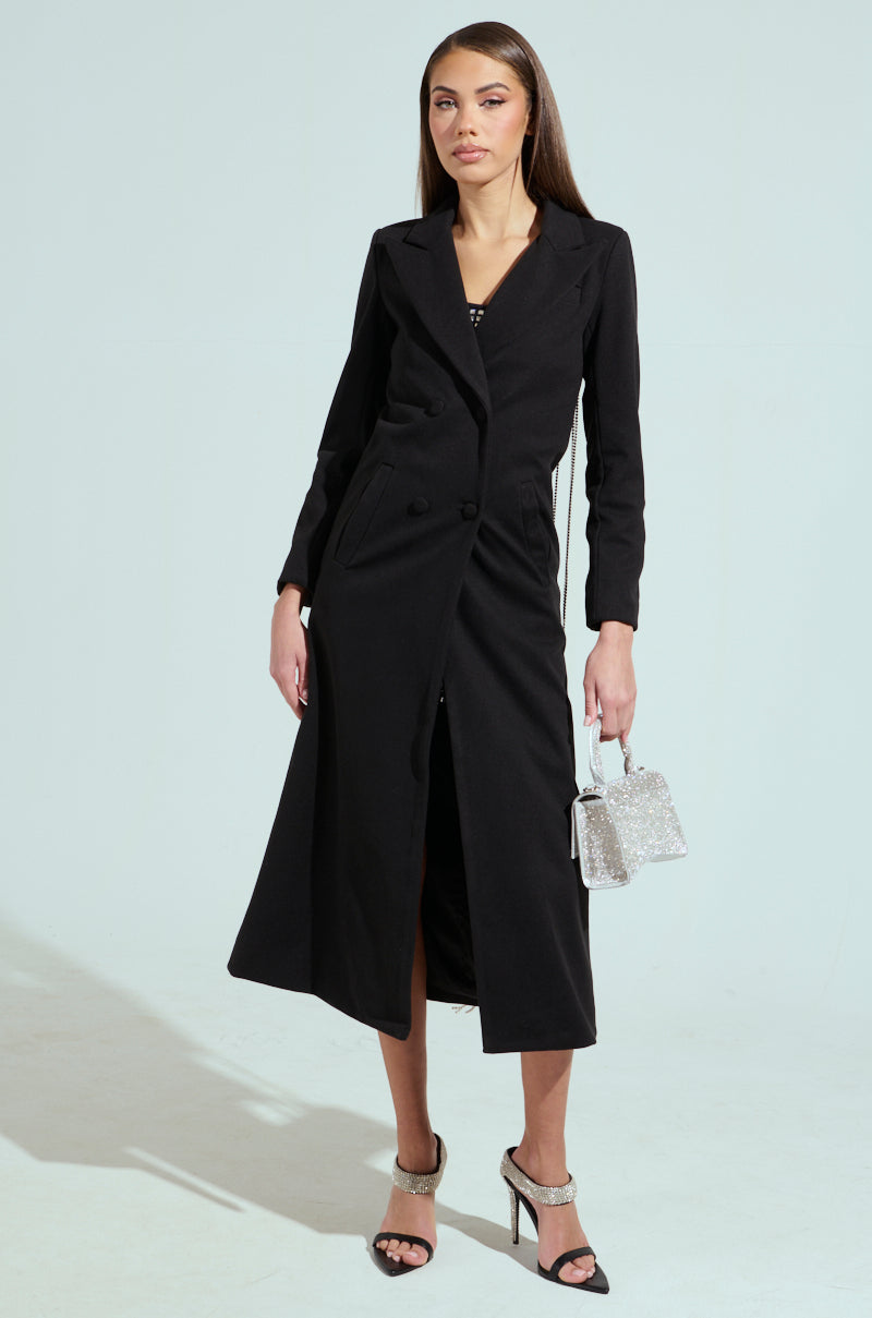 A person stands confidently in the PARTY IN THE BACK RHINESTONE FRINGE TRENCH, a long black coat with a notch lapel collar and high heels. They hold a small sparkling handbag, while the minimalist, light background enhances the chic longline silhouette of their stylish attire.
