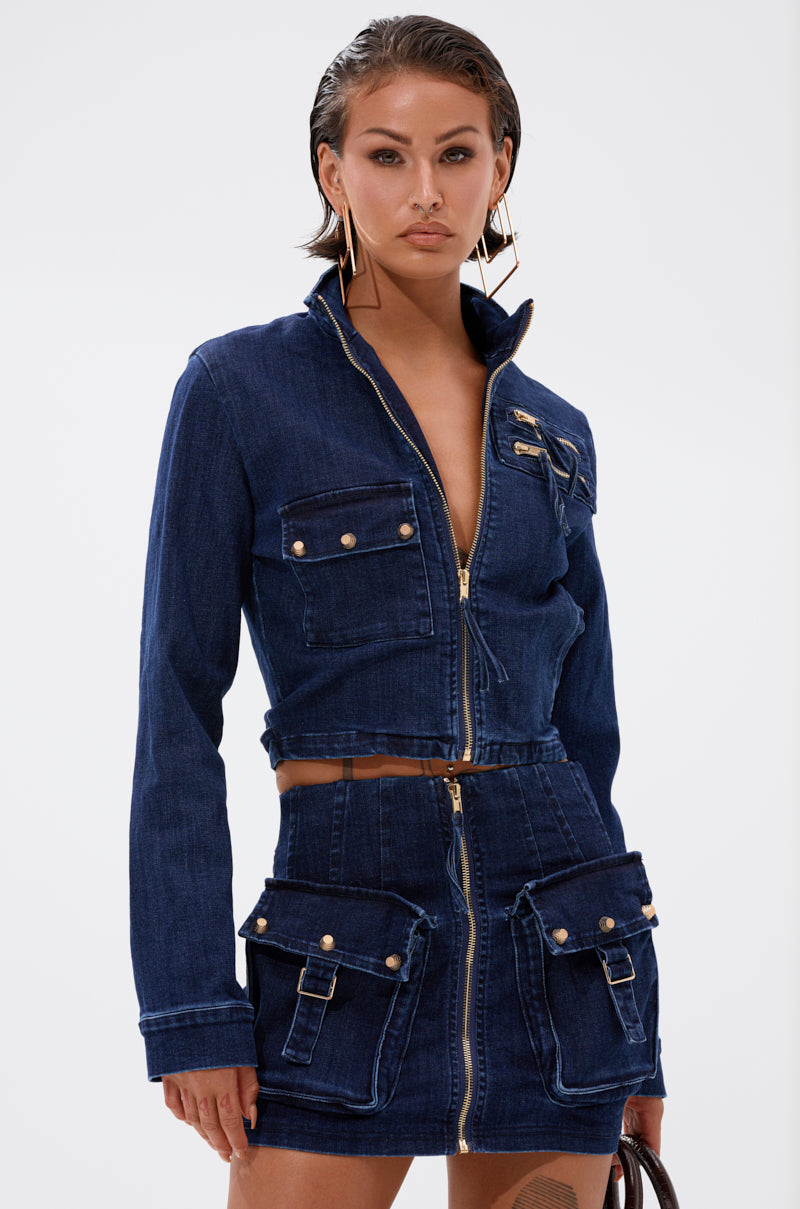 A woman wears the dark blue SOMETHING BETTER DENIM ZIP UP TOP and matching mini skirt with gold zippers, button details, and multiple pockets. Her slicked-back wet-look hair complements bold gold geometric earrings. 0