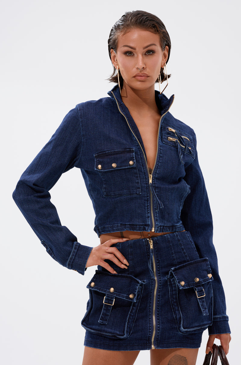 A woman with wet-look slicked-back hair poses confidently with one hand on her hip, wearing the SOMETHING BETTER DENIM ZIP UP TOP in dark blue denim, featuring large front pockets and gold buttons, against a plain background. 1