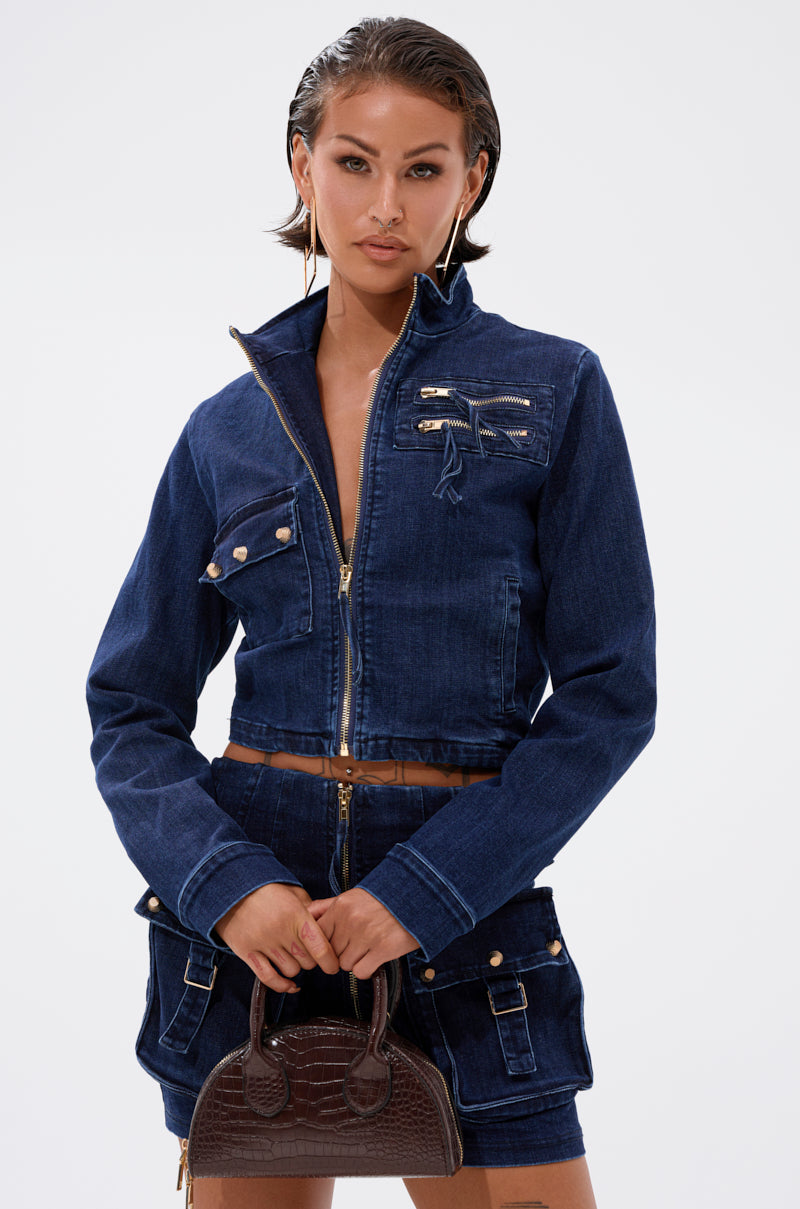 A woman in the SOMETHING BETTER DENIM ZIP UP TOP and matching skirt with gold zippers holds a small brown textured handbag. She has slicked-back hair and gold hoop earrings, standing against a plain white background. 2