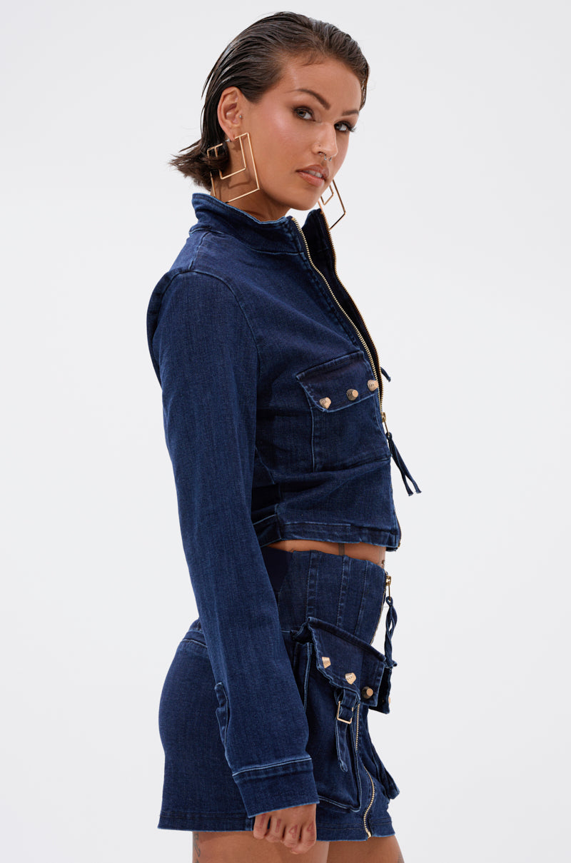 A woman with wet-look hair poses in the SOMETHING BETTER DENIM ZIP UP TOP and a matching high-waisted skirt with metallic buttons, paired with square earrings, against a plain white background. 3