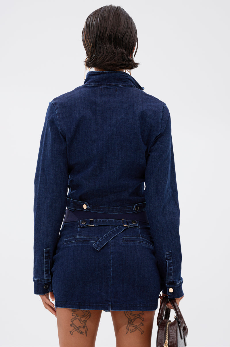 A person with short, dark, wet-look hair is shown from the back wearing the SOMETHING BETTER DENIM ZIP UP TOP and matching skirt. They have lace-up tattoo designs on their thighs and are holding a brown handbag. 4