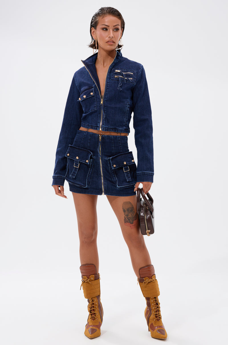 A woman in the SOMETHING BETTER DENIM ZIP UP TOP and a matching mini skirt with multiple pockets, tan lace-up boots, and a small handbag stands against a plain white background. 6