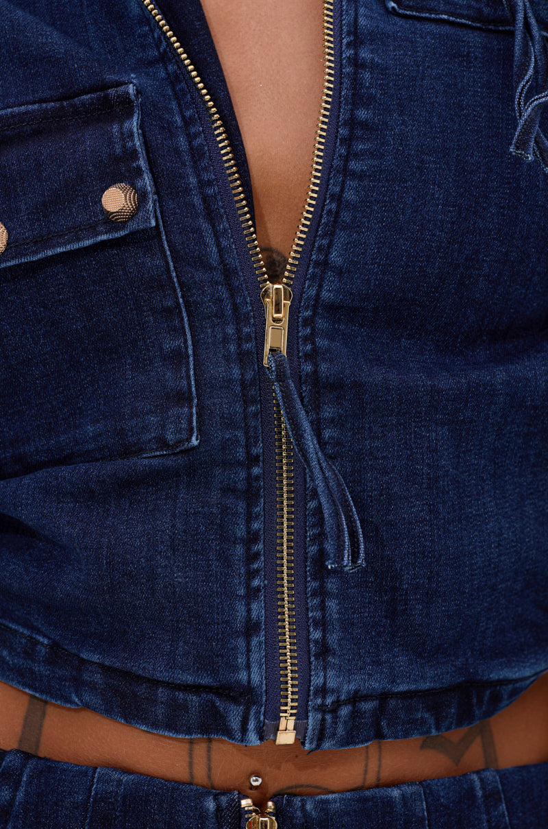 Close-up of a person wearing the SOMETHING BETTER DENIM ZIP UP TOP, a cropped jacket with brass buttons, partially unzipped to reveal skin and a belly piercing above matching denim pants. 8