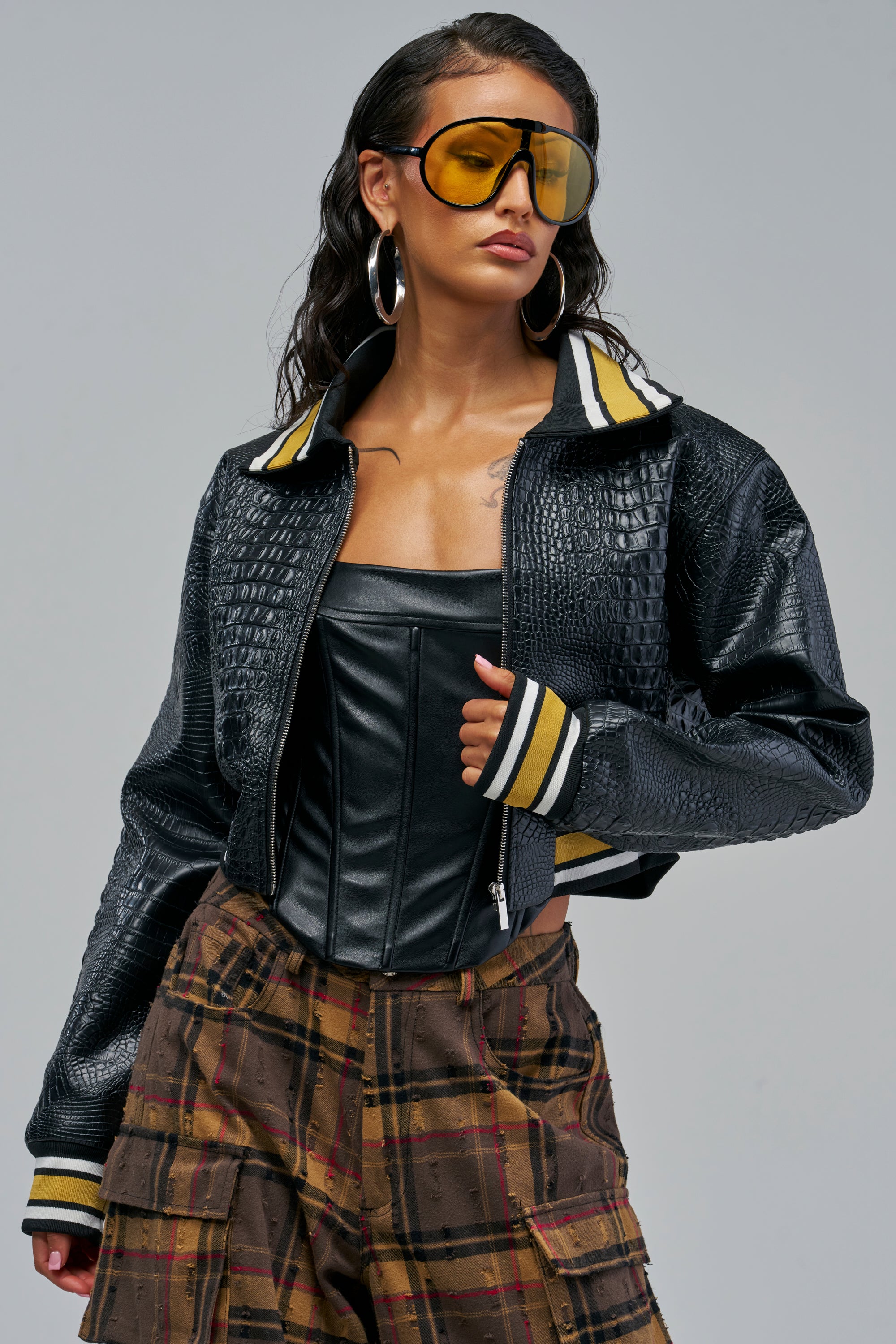 A person wearing a WHAT YOU KNOW CROC BOMBER in black crocodile-embossed leather with striped cuffs and collar over a black corset top, paired with brown plaid cargo pants and yellow-tinted shield sunglasses, showcasing modern streetwear layering.