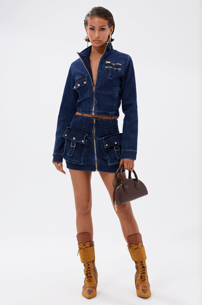 A woman stands confidently in a dark denim zip-up jacket and the SOMETHING BETTER DENIM MINI SKIRT with cargo pockets, tan lace-up boots, thick socks, and a small brown handbag against a plain white background. 0