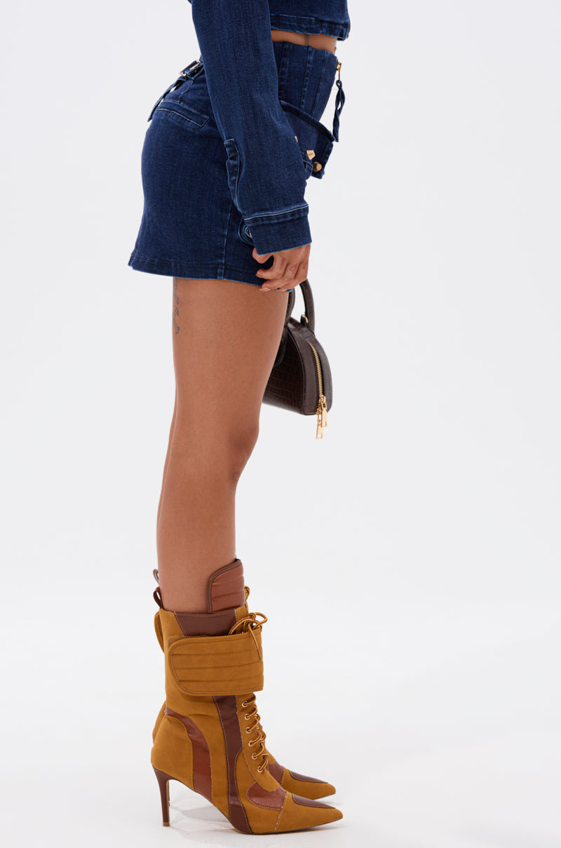 A person stands in profile wearing the SOMETHING BETTER DENIM MINI SKIRT, a cropped denim jacket, brown and mustard yellow heeled boots, and holding a small dark handbag against a plain white background. 3