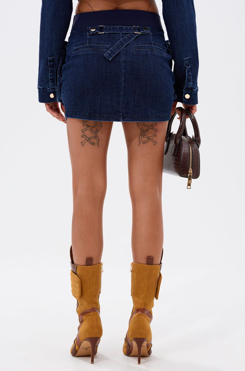 A person wearing the SOMETHING BETTER DENIM MINI SKIRT, a dark denim jacket, tan heeled boots, and holding a brown handbag. Shot from behind, their thigh tattoos are visible. 4