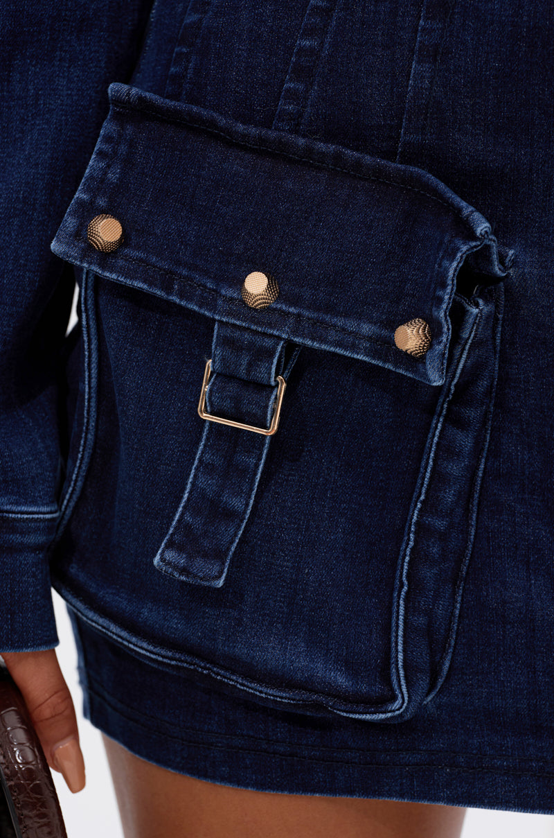Close-up of the SOMETHING BETTER DENIM MINI SKIRT in dark blue, showcasing a large flap pocket with three metallic buttons, buckle detail, high-waisted fit, and part of a hand visible at the hem. 7