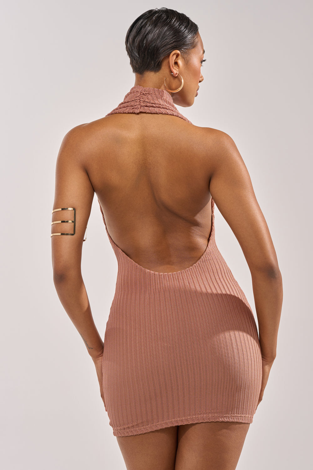 A woman with short hair is shown from the back wearing the HEARTBREAKER WOVEN MINI DRESS IN BROWN, a sleeveless ribbed halter mini dress, along with a three-bar gold arm cuff, against a plain light background. 3