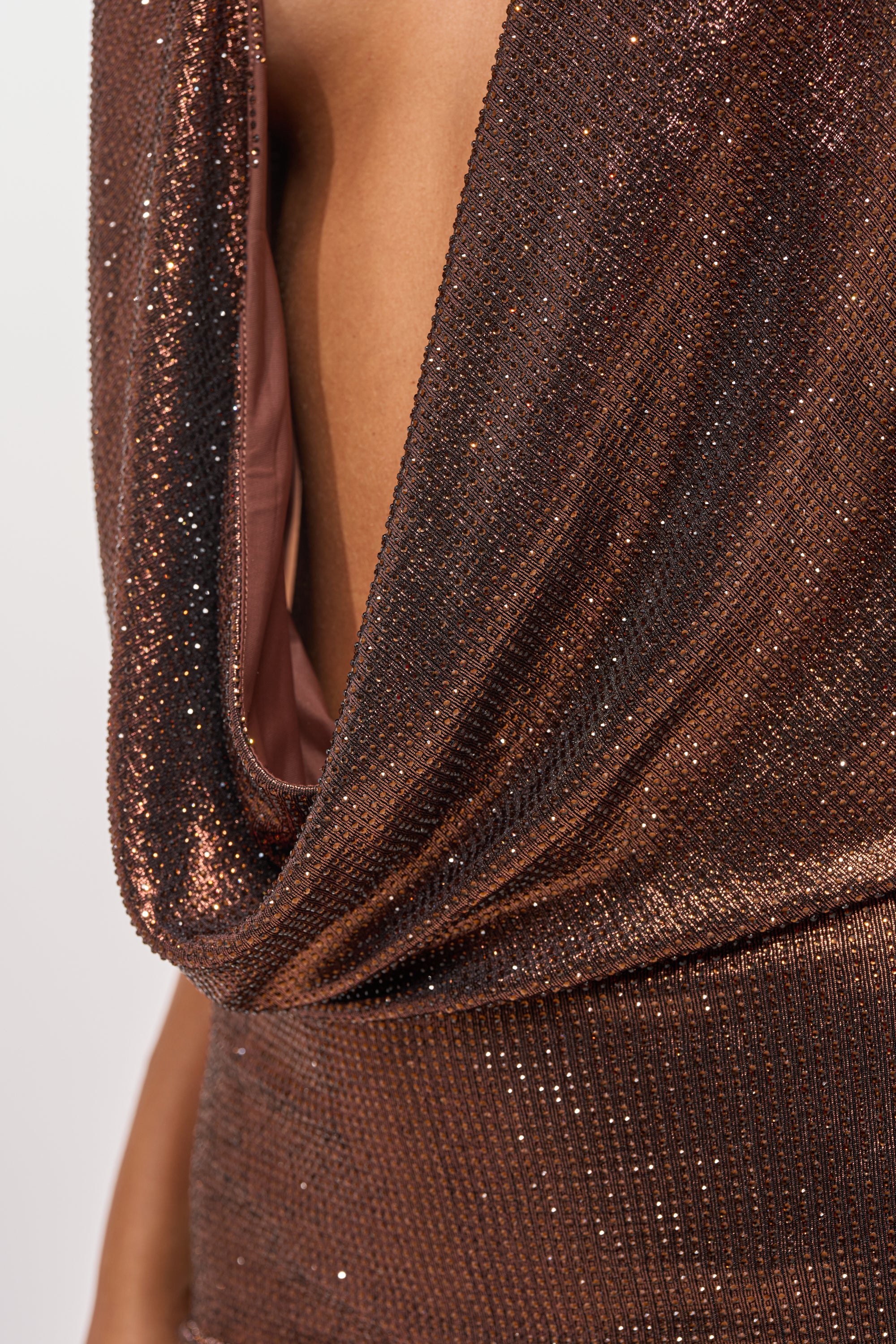Close-up of a person wearing the COME AND GET IT RHINESTONE MINI DRESS IN CHOCOLATE, featuring a shimmery metallic brown fabric and deep draped cowl neckline; only part of the torso and shoulder are visible as the dress sparkles under light. 9