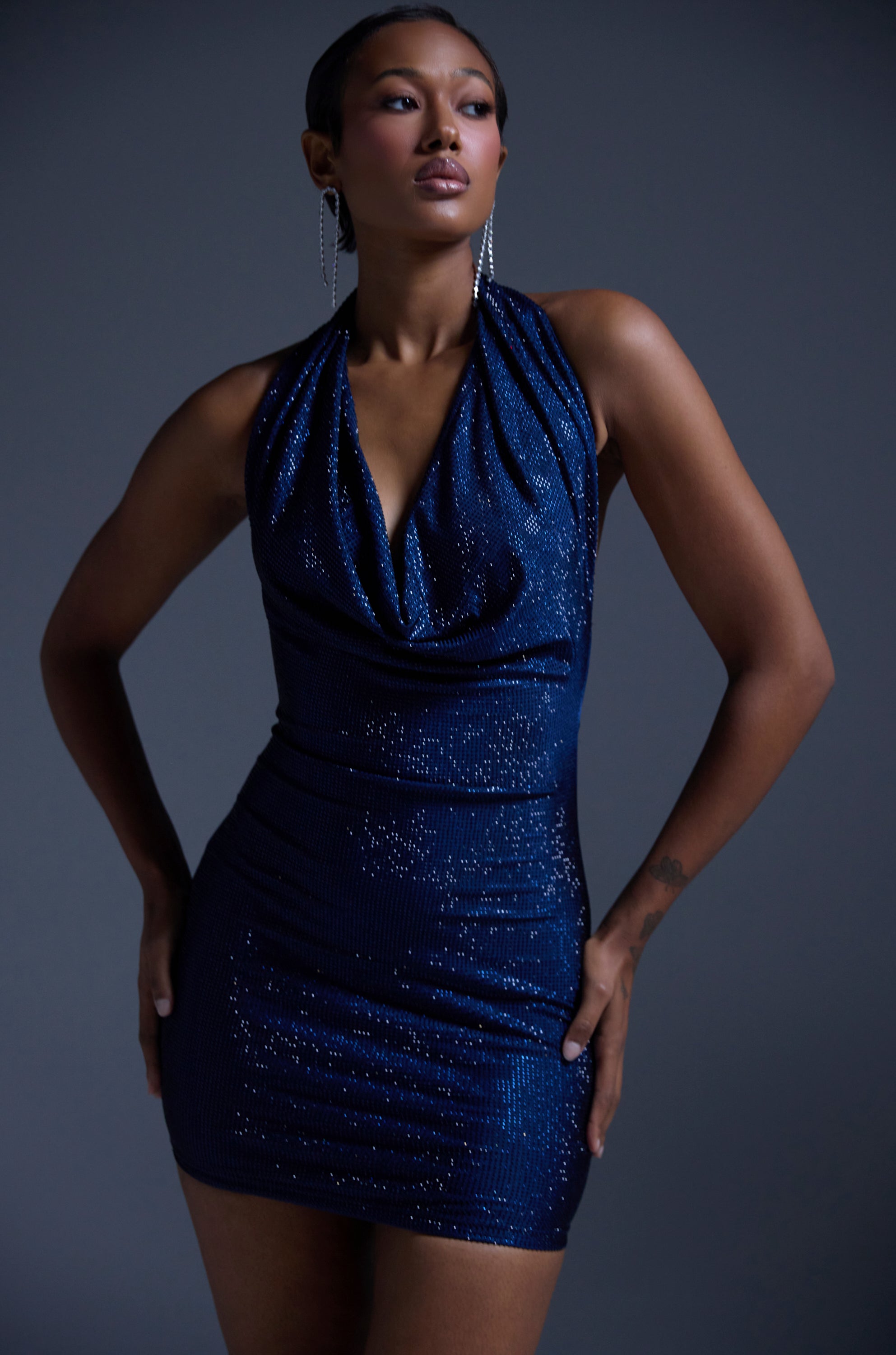 A woman exudes confidence in the HEARTBREAKER RHINESTONE MINI DRESS IN NAVY, featuring a draped neckline and sparkly finish. Posing against a gray background with long earrings and chic short hair, she’s ready for any party occasion.