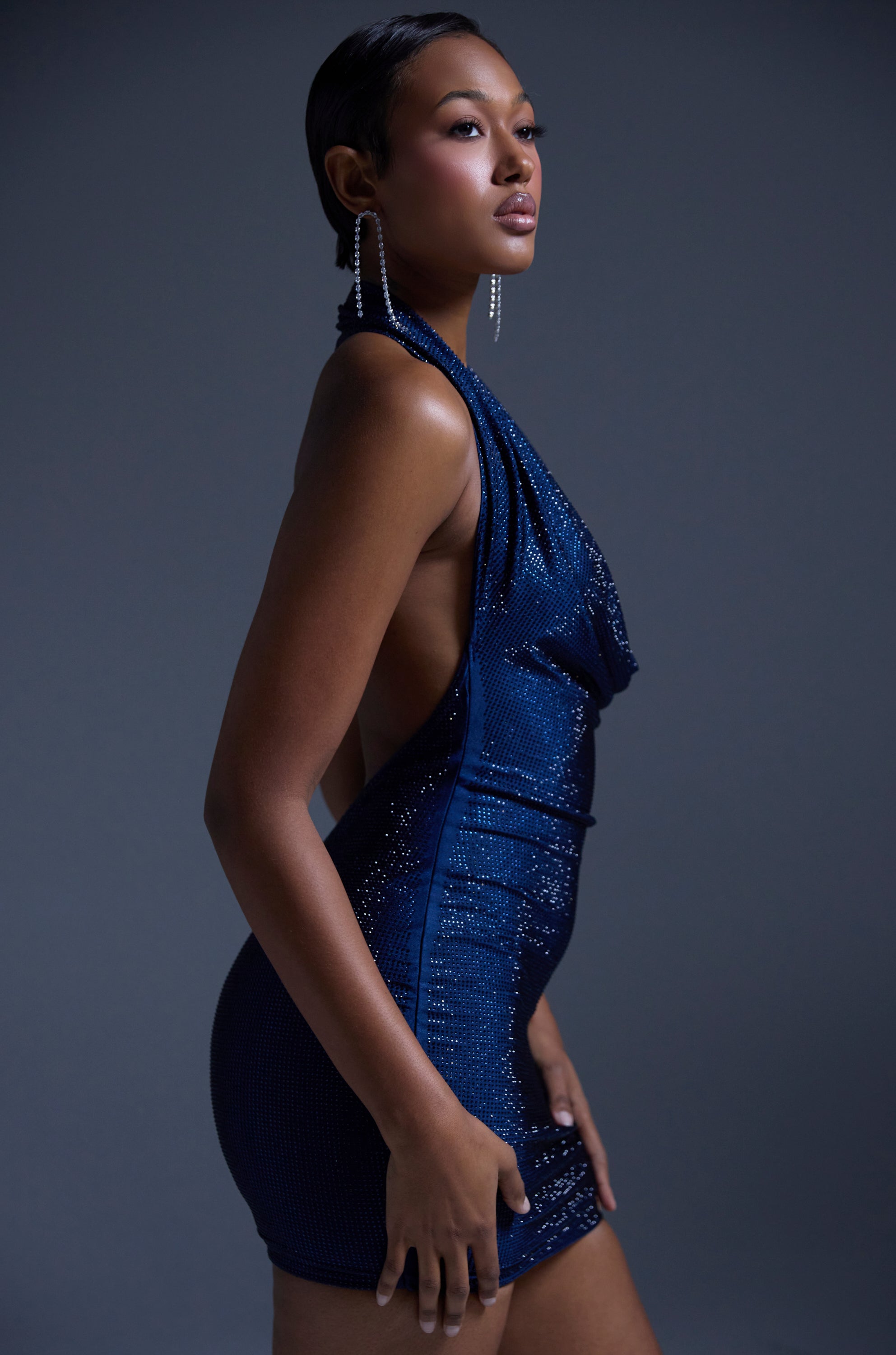 A woman poses confidently in the HEARTBREAKER RHINESTONE MINI DRESS IN NAVY, a sleeveless party dress with open sides, paired with long, dangling earrings, against a plain dark gray background.