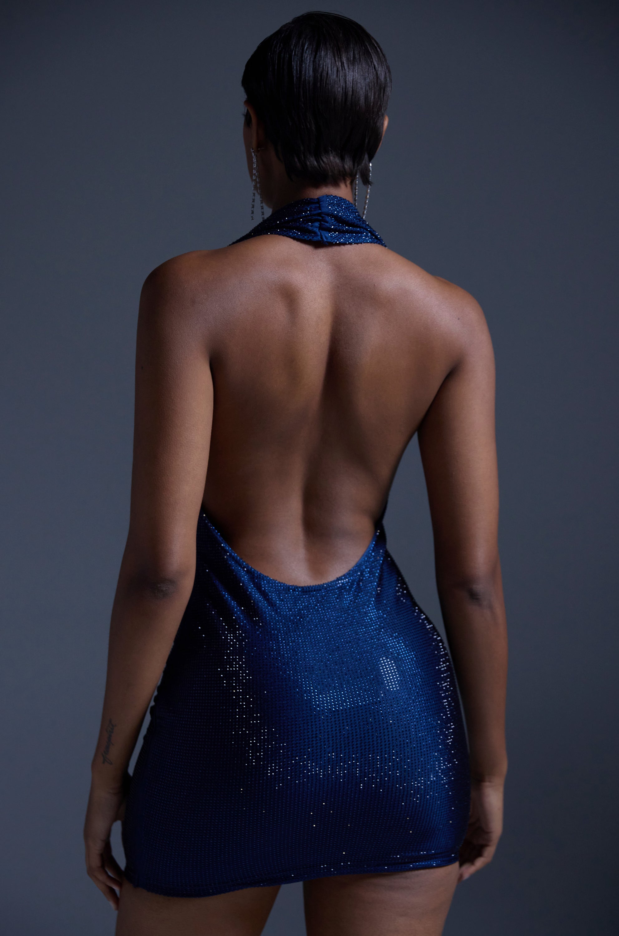 A person with short hair is shown from the back, wearing the HEARTBREAKER RHINESTONE MINI DRESS IN NAVY featuring a sparkling halter neckline and open-back design, set against a plain dark background.