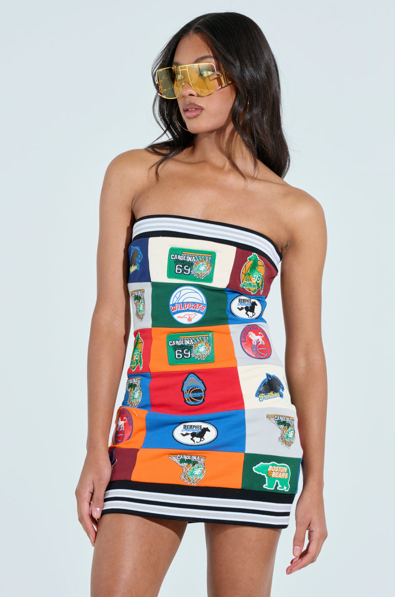 A woman confidently poses in an ALL AROUND THE WORLD PATCHWORK MINI DRESS, a strapless bodycon mini adorned with logos and emblems. She accessorizes with large yellow-tinted sunglasses, her hair flowing freely against a plain white background, highlighting her vibrant style.