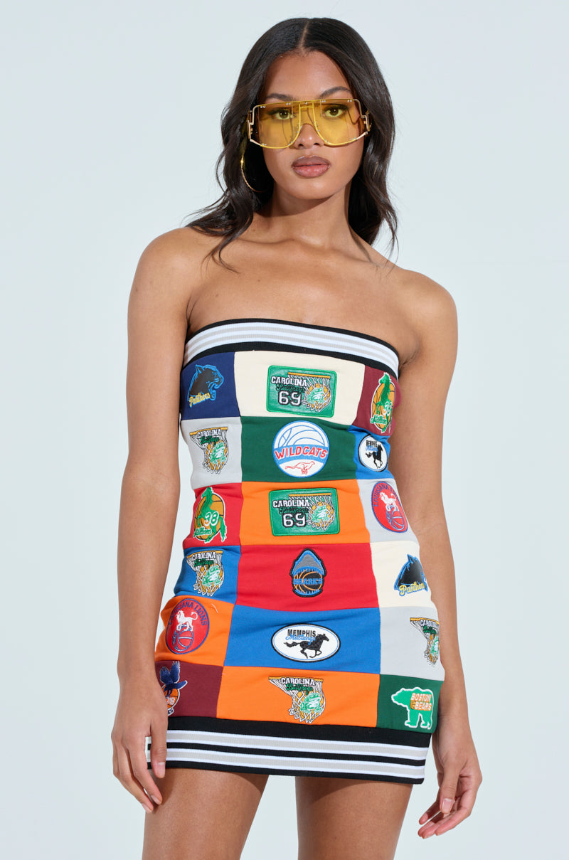 Against a plain background, a confident woman sports yellow sunglasses and the ALL AROUND THE WORLD PATCHWORK MINI DRESS, showcasing its vibrant colorblock design with various logos. Her long hair cascades down, complementing the bold bodycon silhouette.