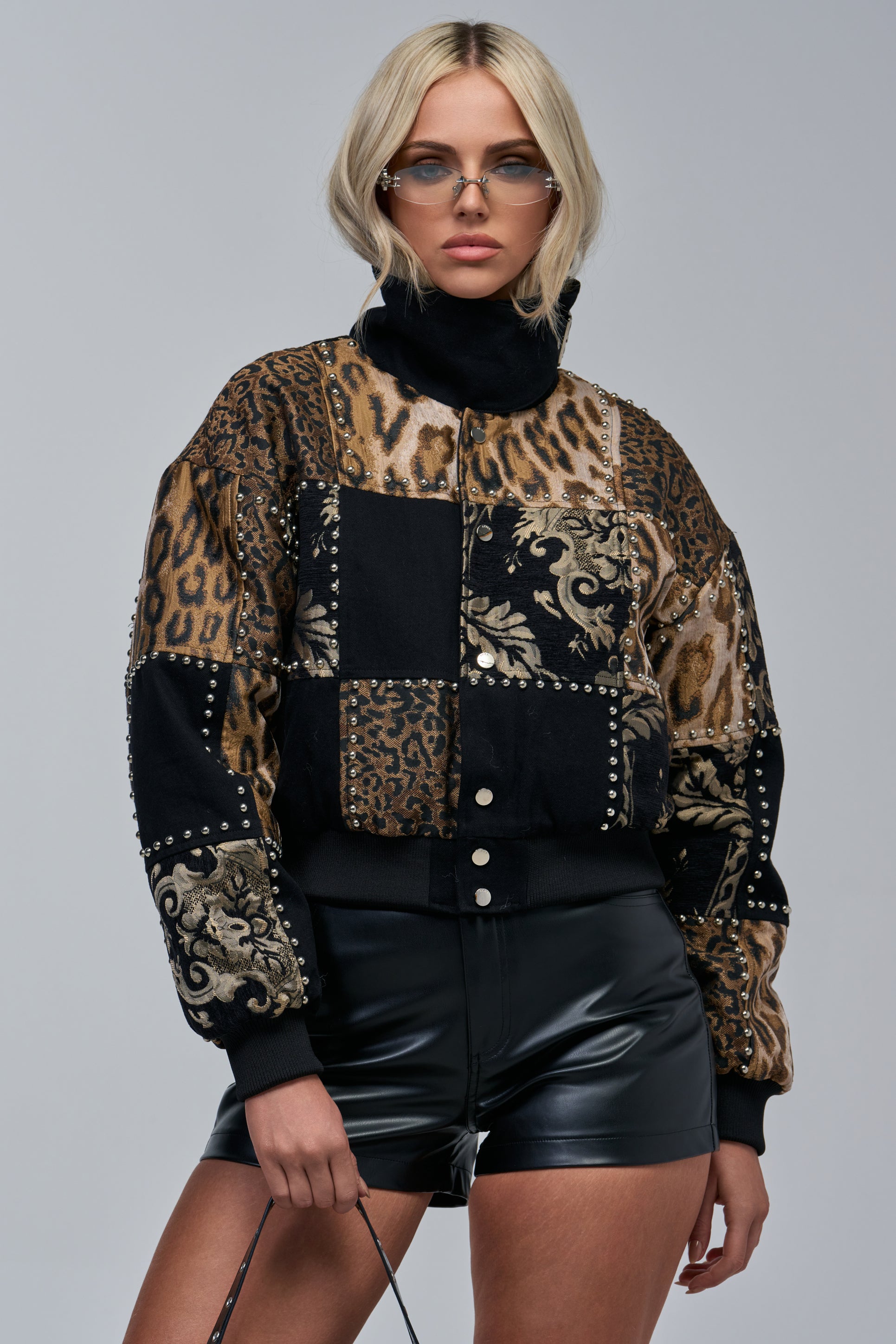 A woman wearing sunglasses and the HEX PATCHWORK BOMBER—featuring animal print and baroque patterns with metal studs—paired with black leather shorts, stands against a plain gray background.