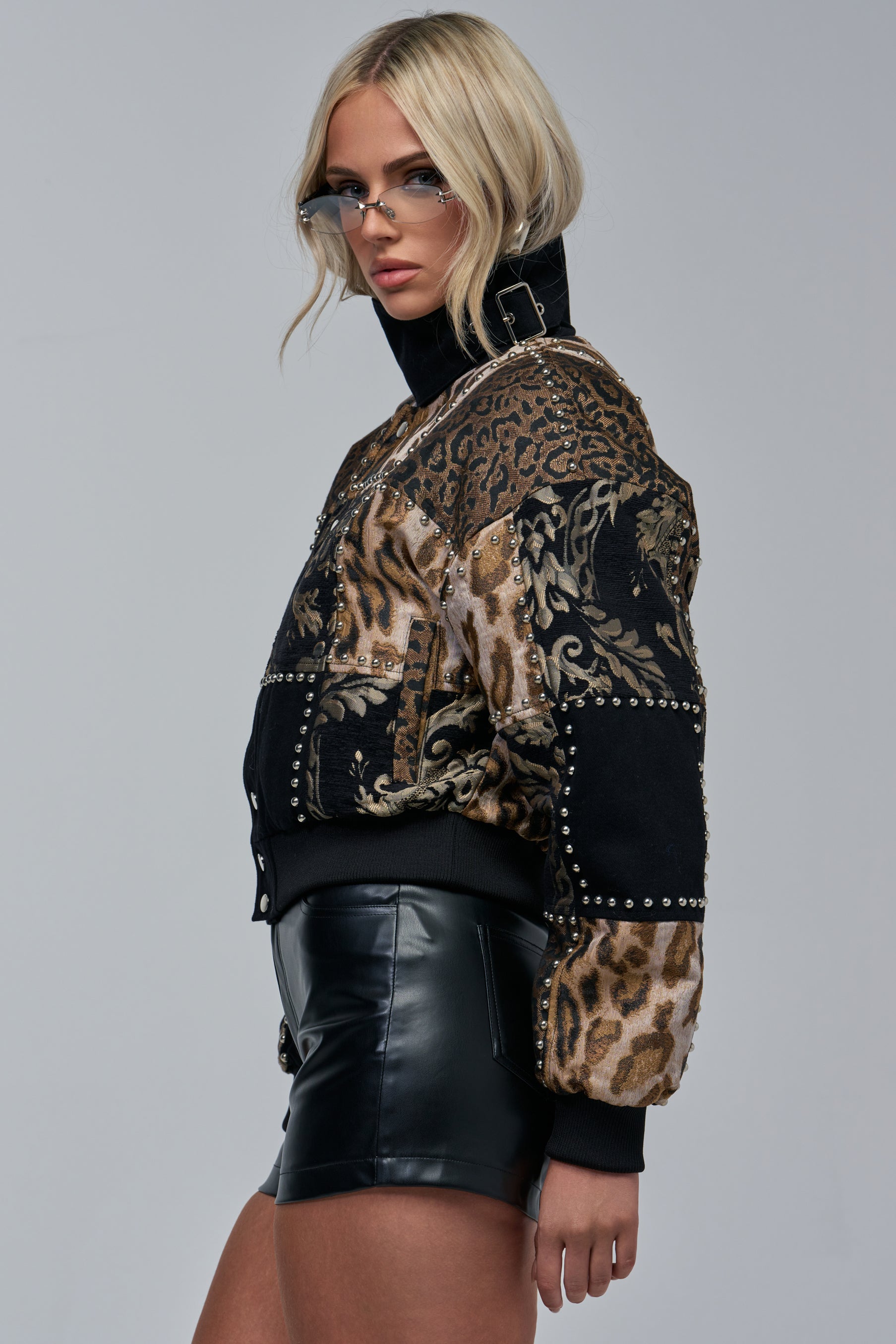 Woman with blonde hair wearing the HEX PATCHWORK BOMBER, black leather shorts, and clear glasses poses sideways against a plain grey background.