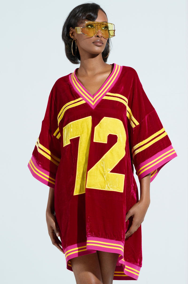 A woman wears the MOVING FORWARD VELVET JERSEY DRESS, a red oversized jersey with pink and yellow varsity stripes and a bold yellow "72" on the front. She accessorizes with yellow-tinted sunglasses, hoop earrings, and has shoulder-length hair against a light background. 0