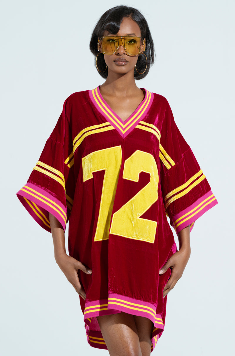 A person in oversized sunglasses and hoop earrings sports the MOVING FORWARD VELVET JERSEY DRESS, featuring a red and gold design with the number 72. The roomy fit includes wide sleeves and bold varsity-inspired yellow and pink stripes, set against a plain light blue background. 1