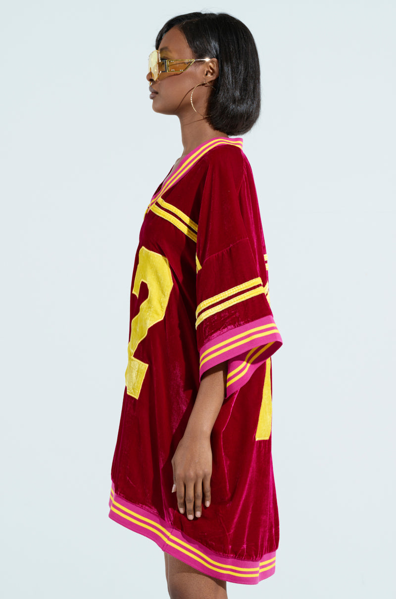 A person in a red MOVING FORWARD VELVET JERSEY DRESS with varsity-inspired stripes and yellow lettering stands in profile against a light background, flaunting shoulder-length hair, large hoop earrings, and sunglasses. 2