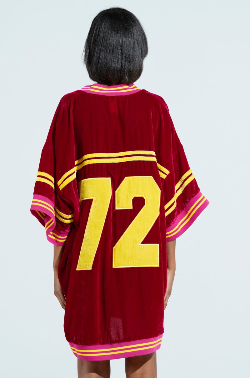 A person with short black hair, wearing an oversized MOVING FORWARD VELVET JERSEY DRESS in red with varsity-style yellow stripes and the number 72 on the back, stands facing away from the camera against a light background. 3