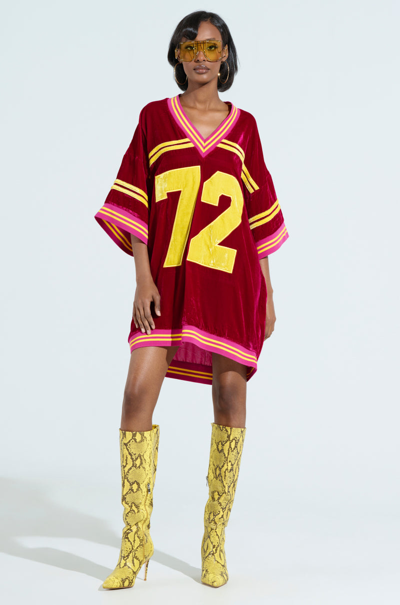 Dressed in the oversized MOVING FORWARD VELVET JERSEY DRESS, with varsity-style sleeve stripes, a prominent yellow "72," and accessorized with yellow sunglasses and knee-high snake-patterned boots. 4