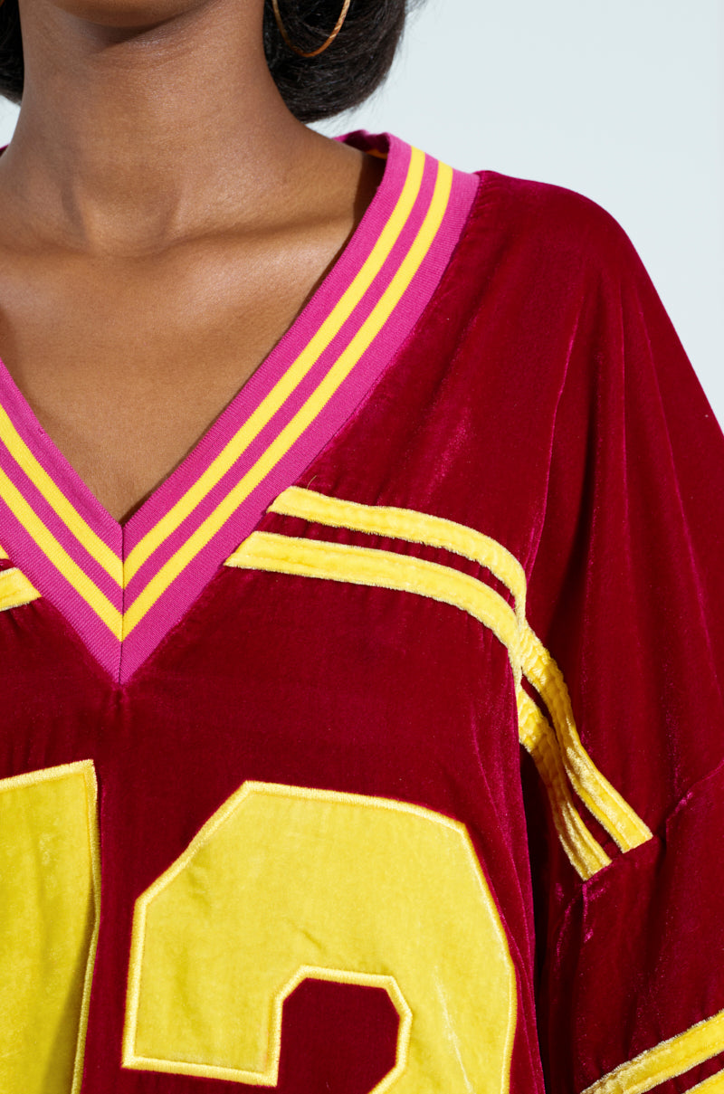 A close-up showcases the MOVING FORWARD VELVET JERSEY DRESS in a red and yellow sports jersey style, with varsity-inspired stripes, a V-neck, and a partially visible "33" on its oversized fit. The light background enhances the vibrant colors of this sporty attire. 7