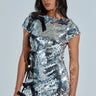 A woman exudes confidence in the "I'M THE GIFT SEQUIN MINI DRESS WITH BOWS," a shimmering silver mini dress with satin accent bows, short sleeves, and a round neckline. Her long dark hair cascades over her shoulders against a neutral background.