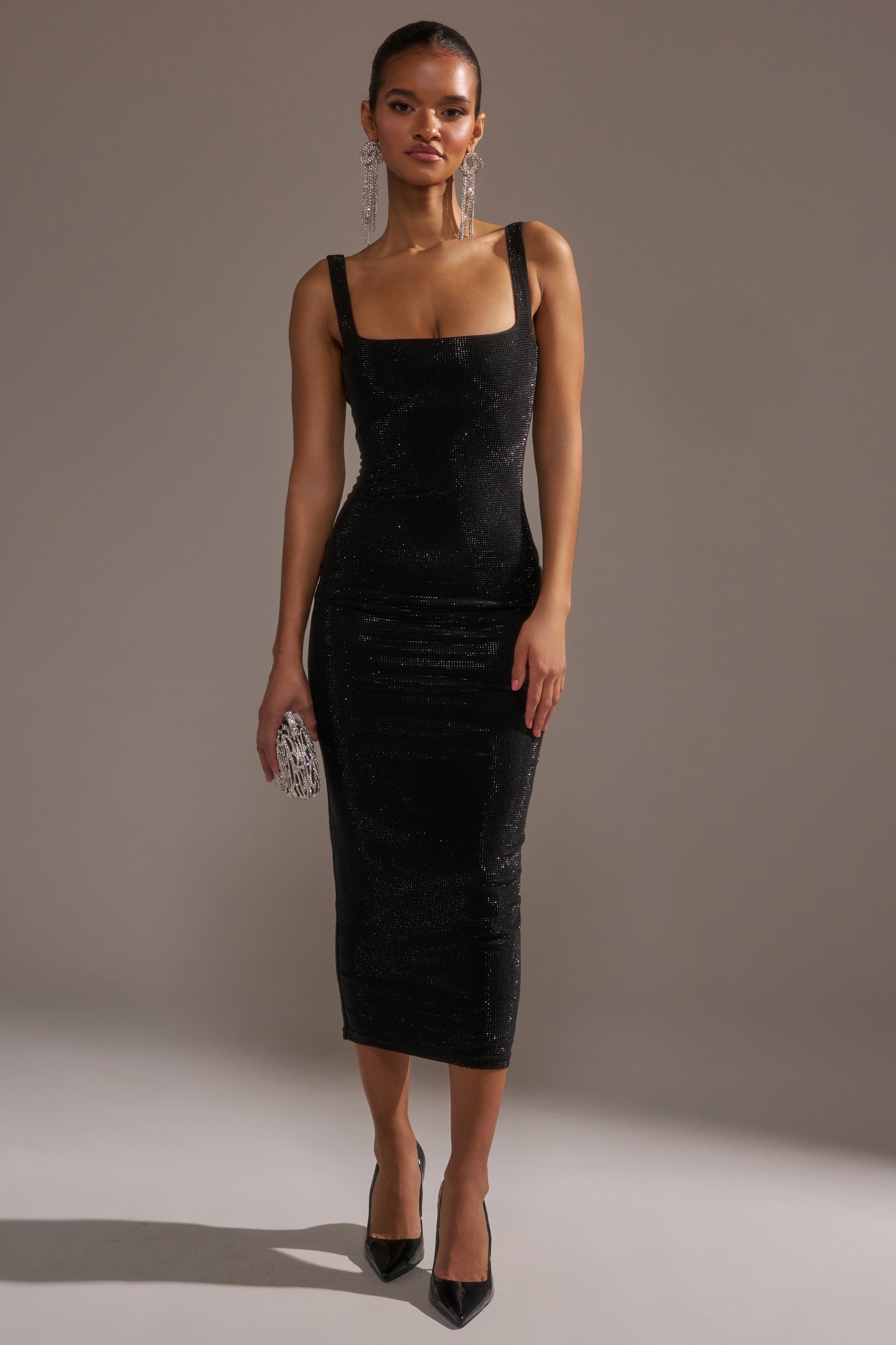 A woman stands against a plain background in the CLASSY BUT SASSY RHINESTONE MIDI DRESS IN BLACK, a form-fitting, sleeveless midi dress with a square neckline. She accessorizes with black heels, statement earrings, and a small silver clutch for an elegant look.