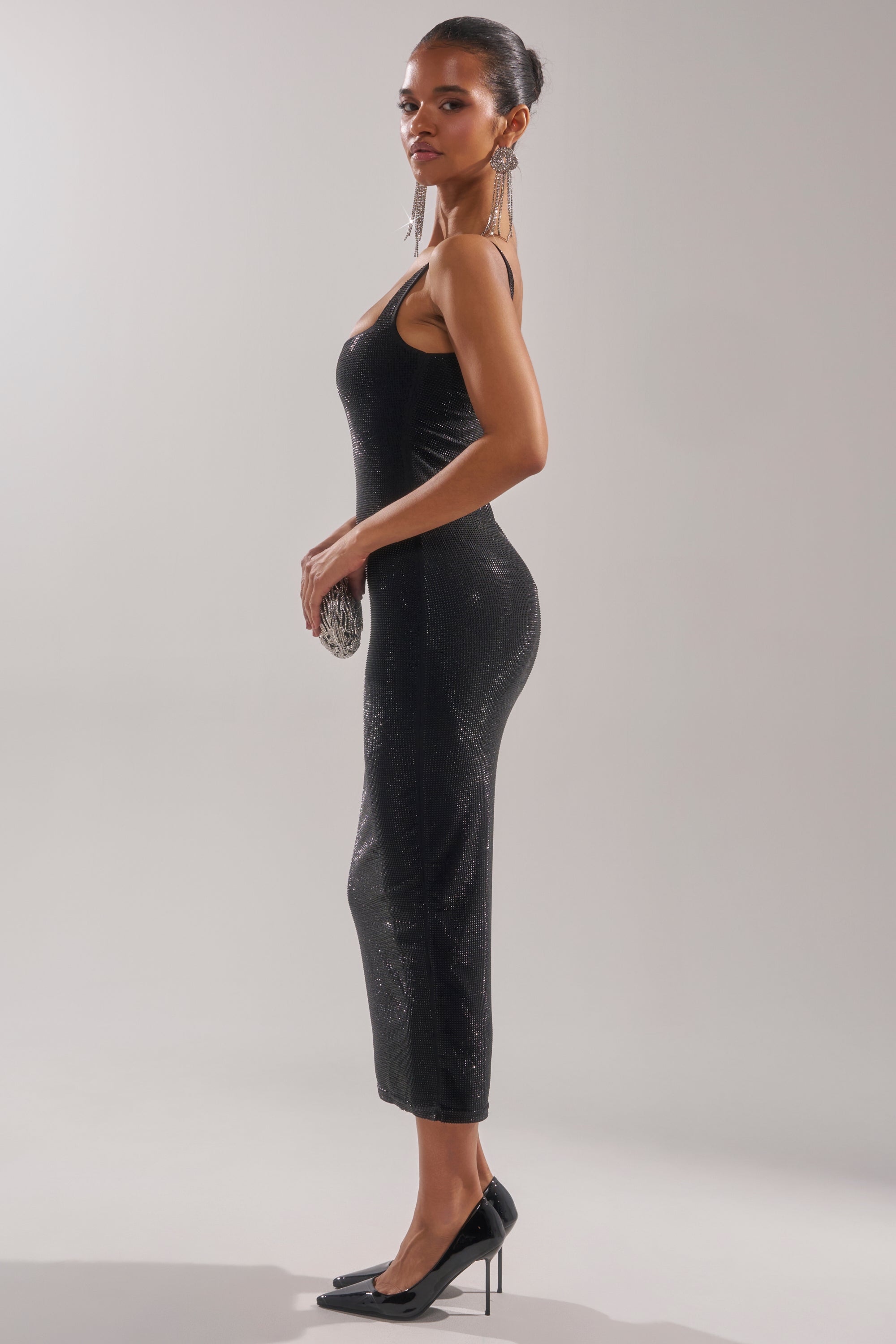 A woman in the CLASSY BUT SASSY RHINESTONE MIDI DRESS IN BLACK and black heels stands in profile, holding a clutch and wearing long earrings. Her hair is styled elegantly against a light background.
