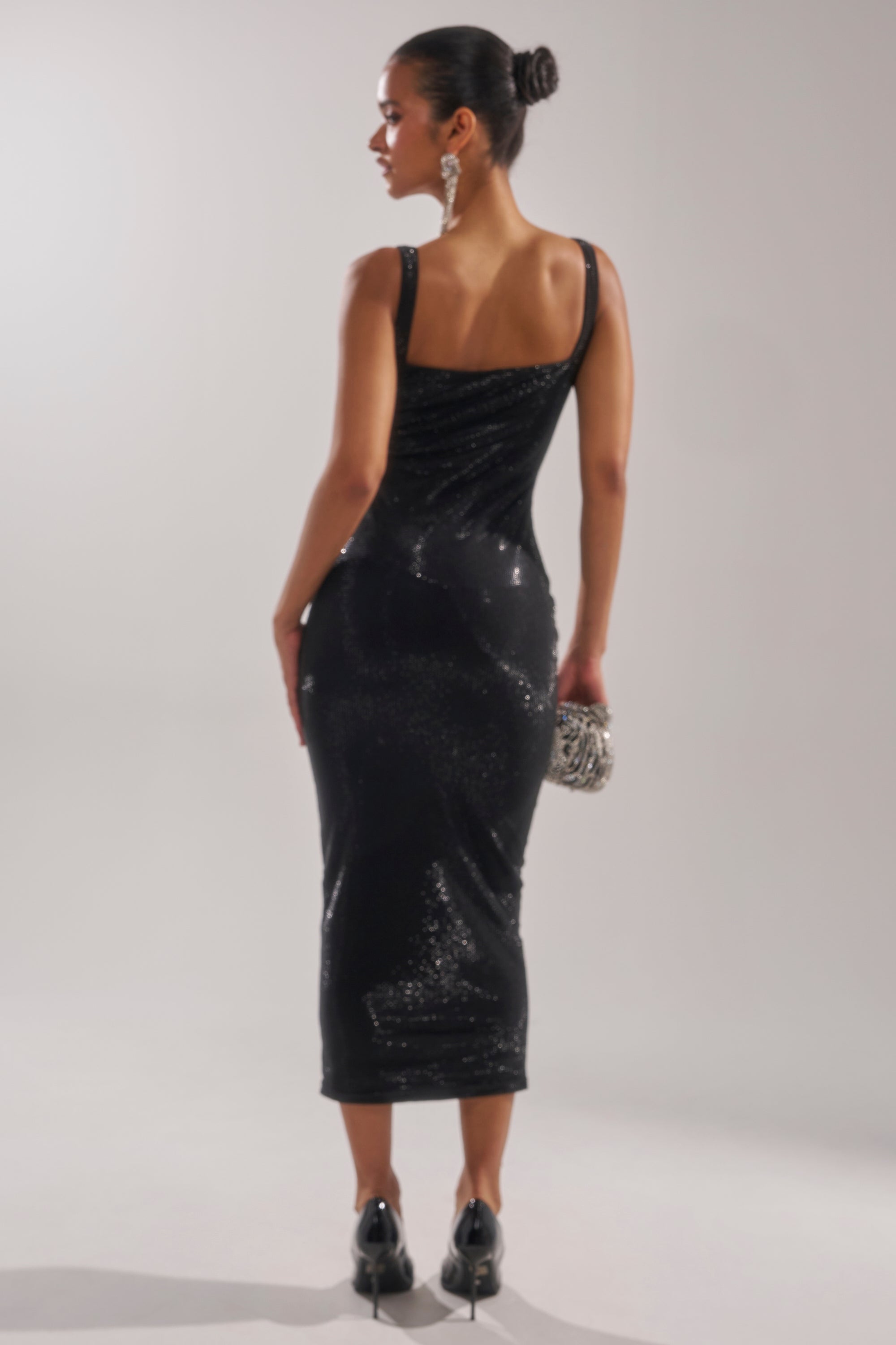 A woman with her hair in a bun stands with her back to the camera, wearing the CLASSY BUT SASSY RHINESTONE MIDI DRESS IN BLACK, black heels, and holding a silver clutch against a plain background.