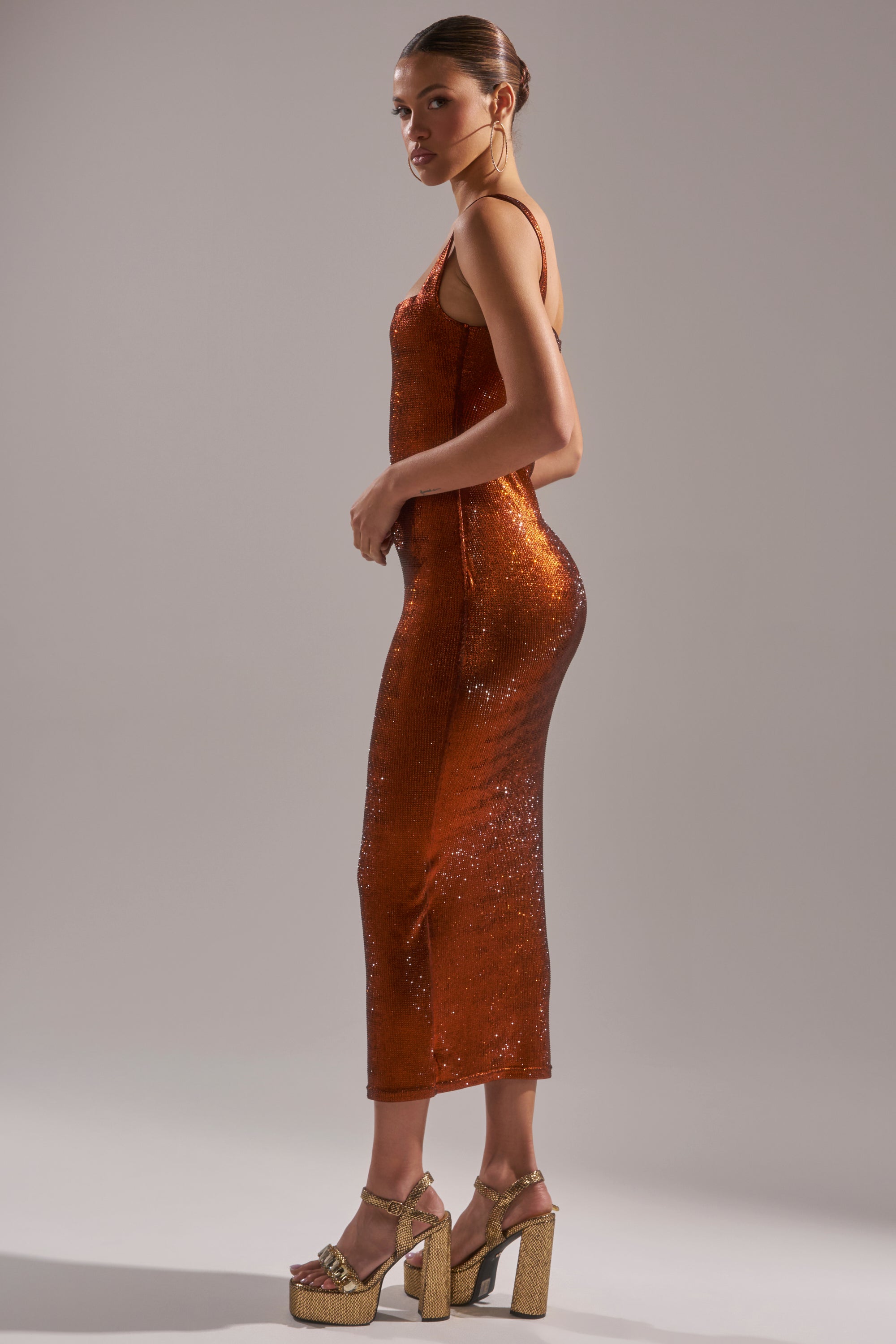A woman poses in the Classy But Sassy Rhinestone Midi Dress in Rust, sleeveless and shimmering, with gold platform heels, standing sideways against a plain light gray background.