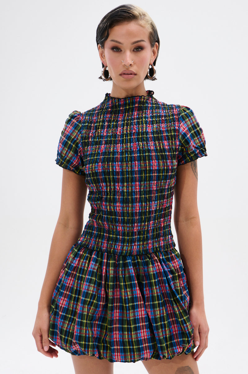 A person with short dark hair models the THAT GIRL PLAID MINI DRESS—a short-sleeved, high-neck mini dress with a smocked bodice and flared bubble skirt—while standing against a plain white background.